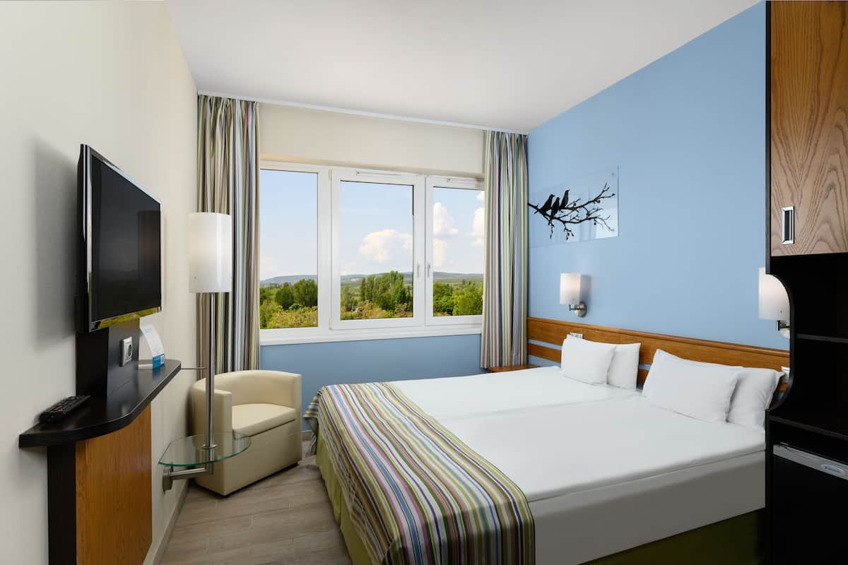 Ein Hotelzimmer mit einem grossen Bett, einem Fernseher, einem Schreibtisch mit Lampe, einem Sessel und einem Fenster mit Blick auf Grünflächen.