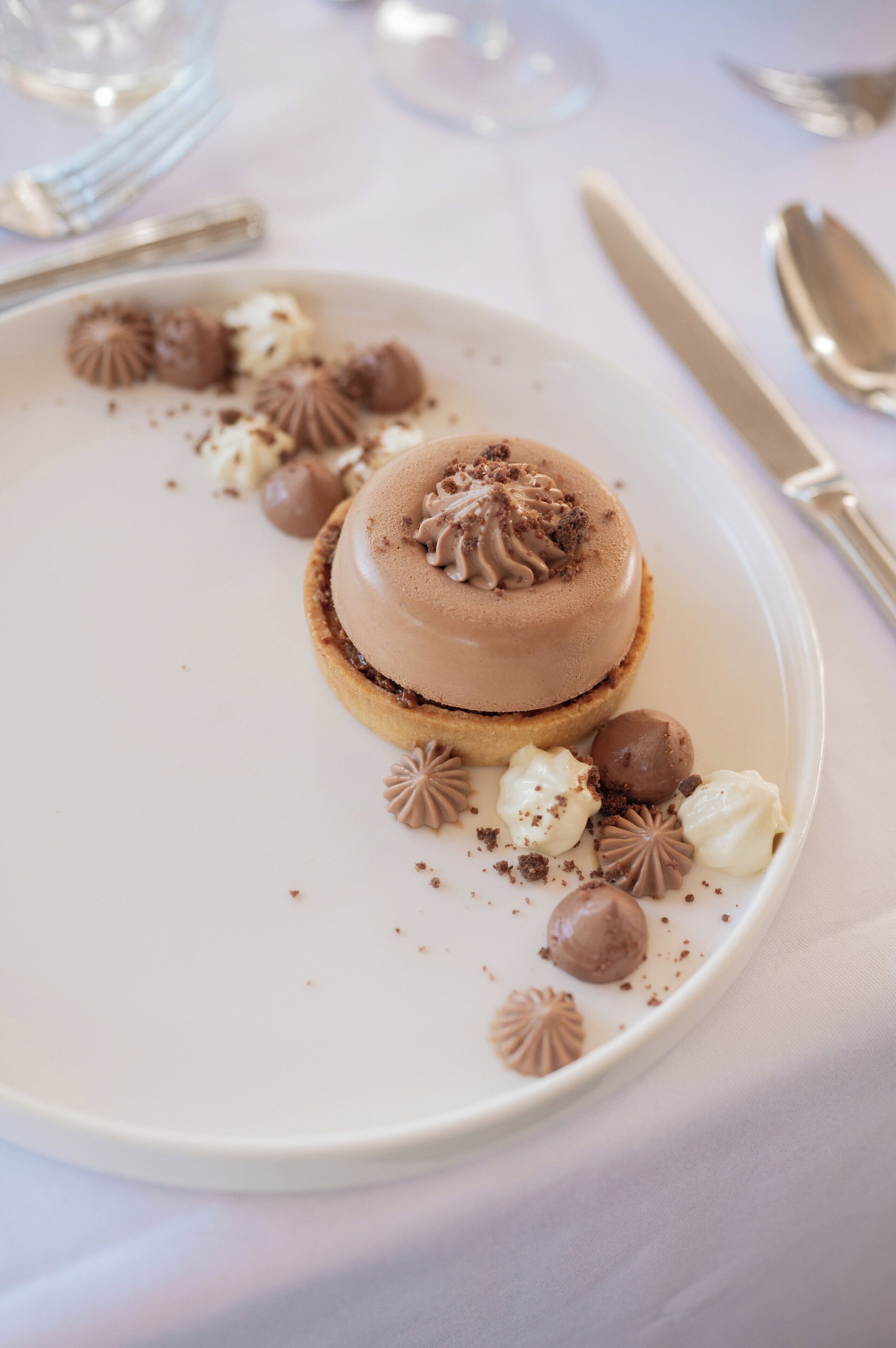 Een nagerecht op een wit bord met chocolade en witte details, met een vork en lepel op een tafel.