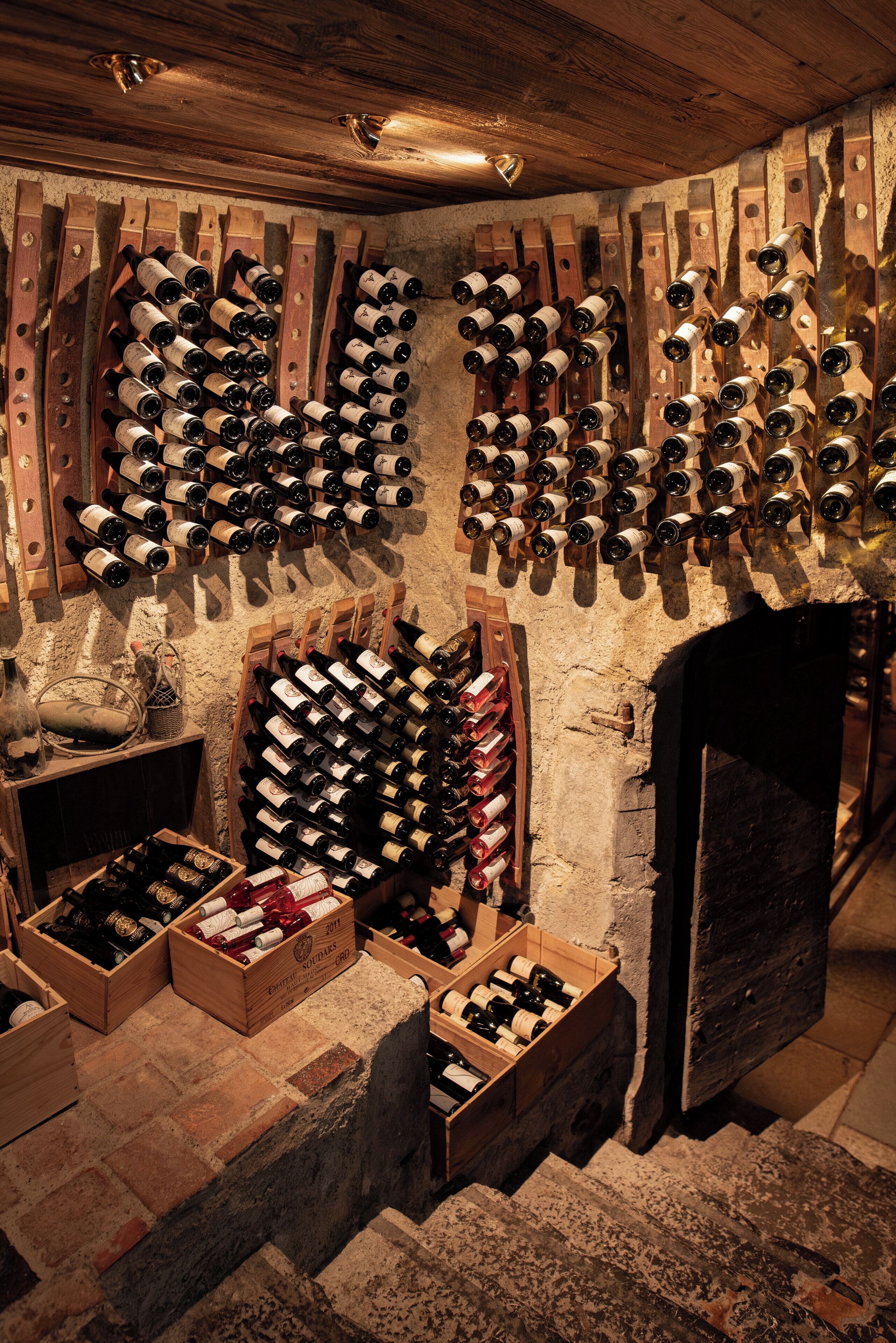 A wine cellar with wooden racks holding numerous bottles, wooden crates with bottles, and a stone wall.