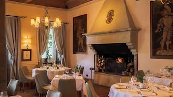 A dining room with round tables, chandeliers, a fireplace, and artwork on the walls.