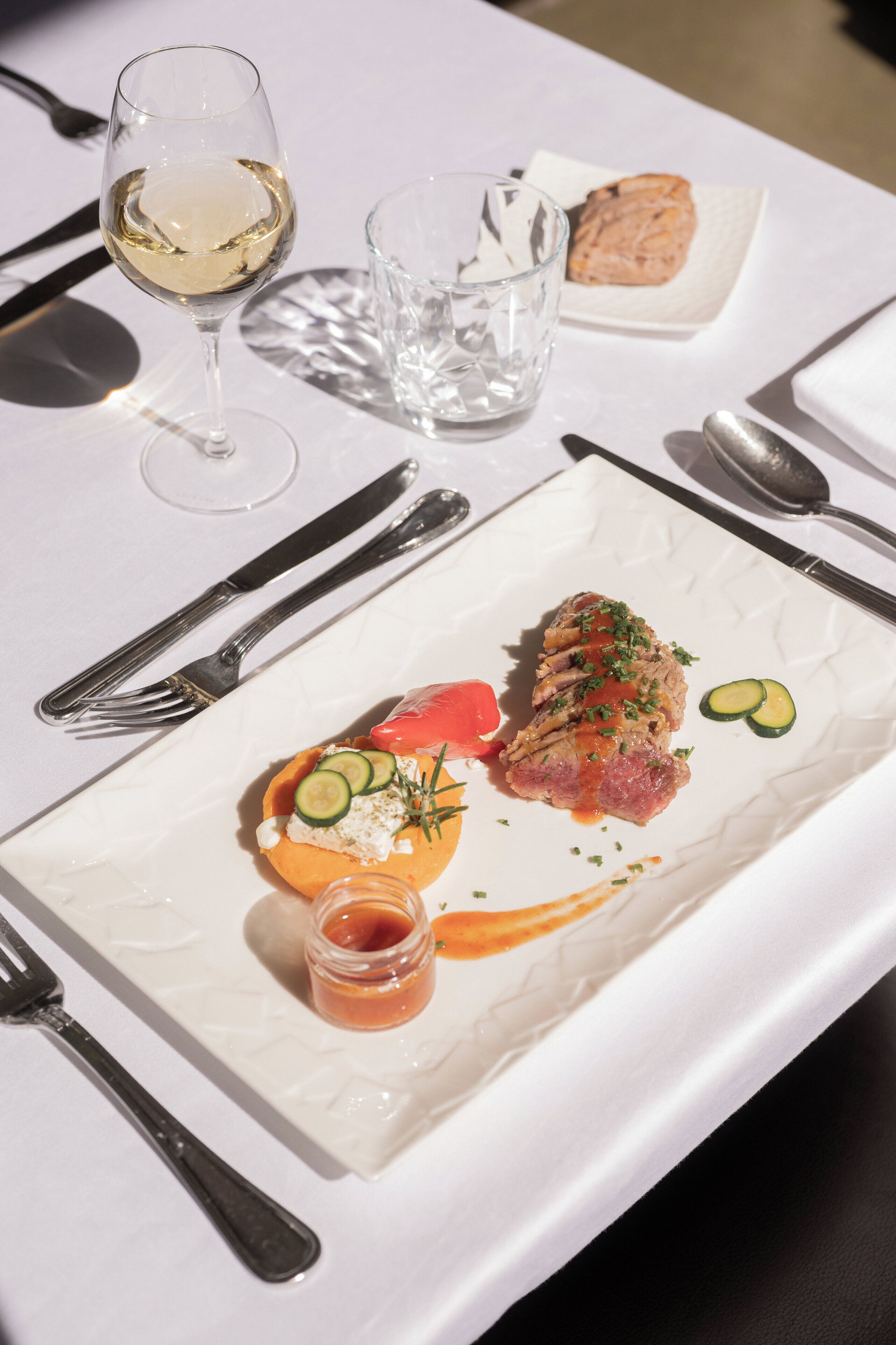 A white plate with a meat dish, a small orange fruit, and a glass of white wine on a table.