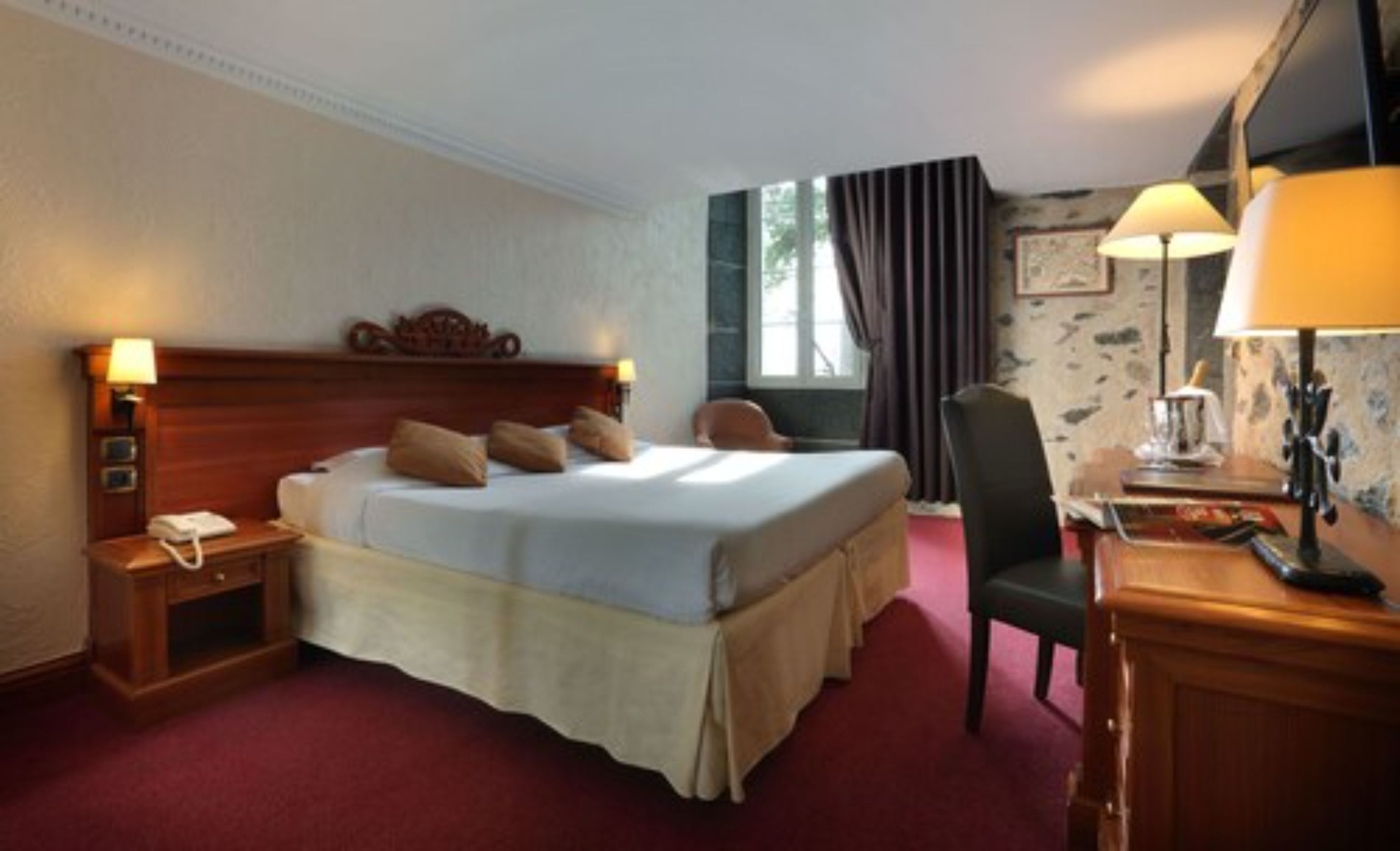 A hotel room with a large bed, a desk with a lamp, a chair, and a telephone.