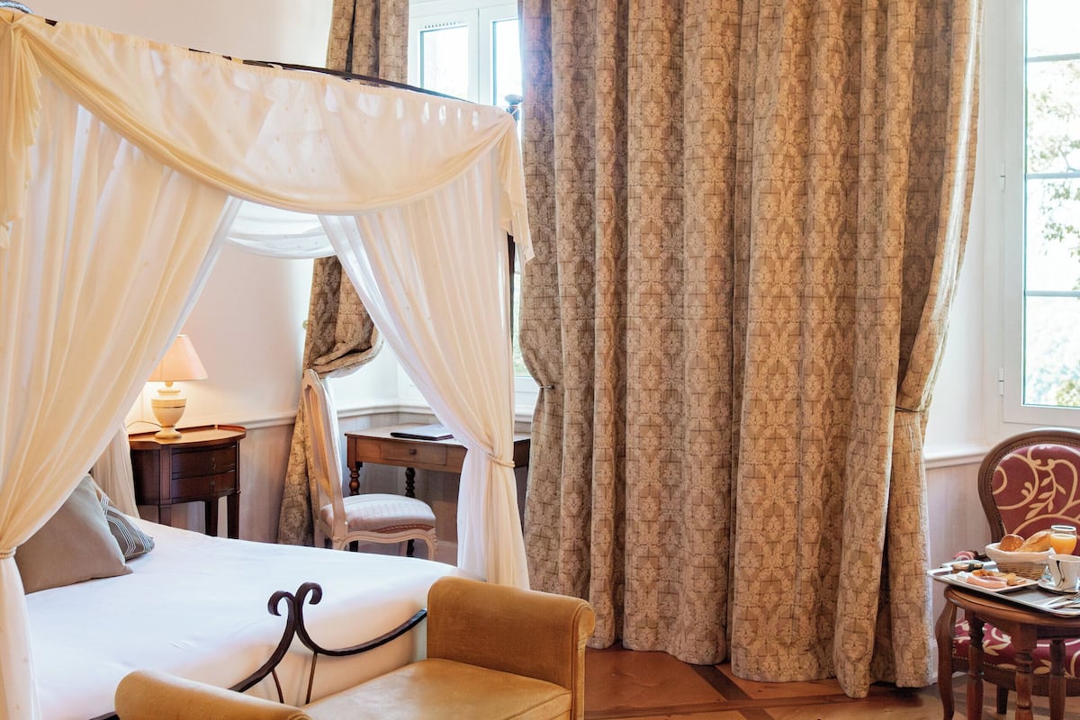 Un dormitorio con una cama a dosel, una silla de madera, una mesita con una bandeja de desayuno y una ventana con cortinas.