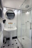 Un baño moderno con un lavamanos pedestal, una ducha con mampara de vidrio y un espejo redondo sobre el lavamanos.