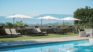 Un espace piscine avec des chaises longues et des parasols, un mur en pierre et une vue sur les montagnes lointaines.