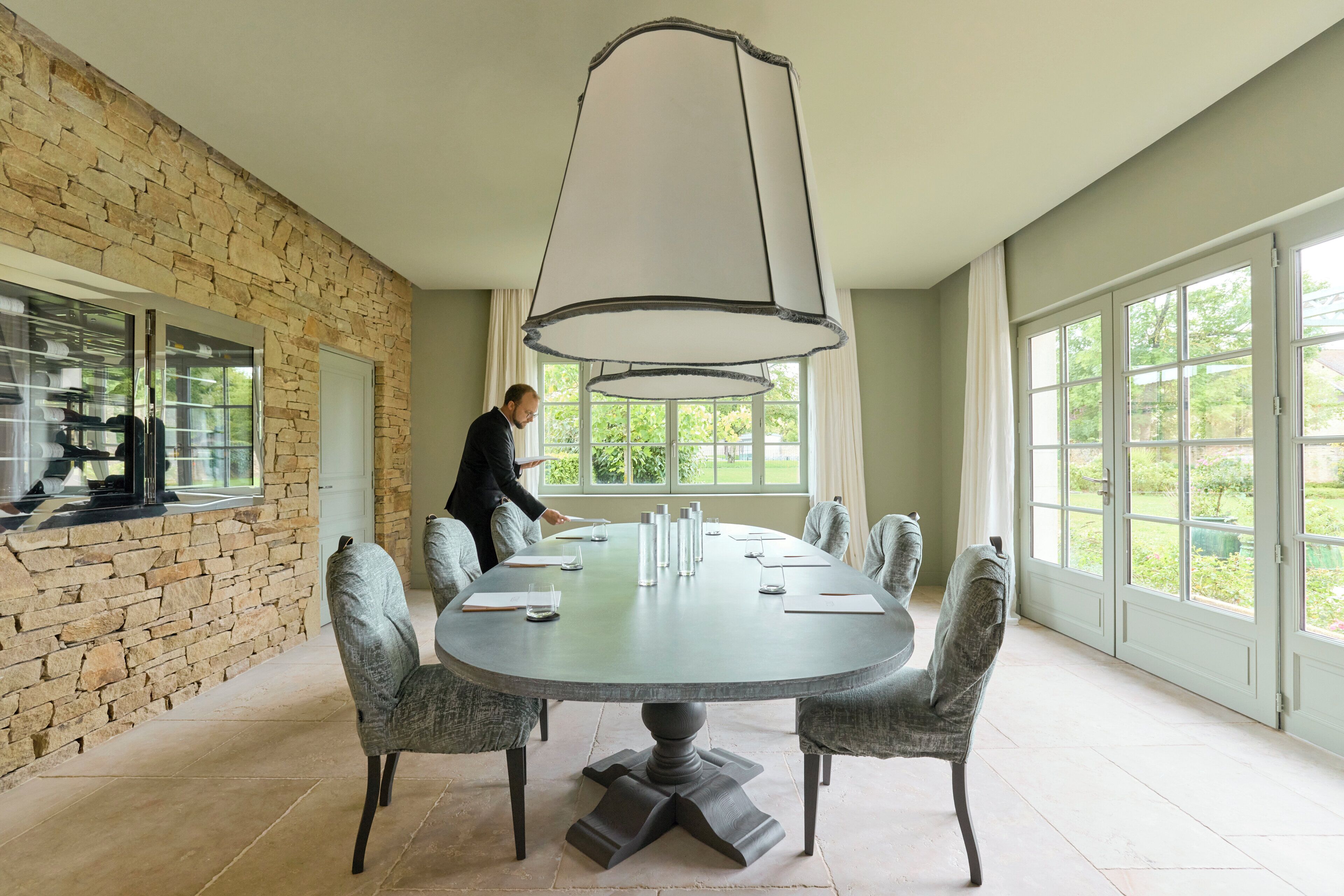 A conference room with a round table, grey upholstered chairs, a large pendant light, and a stone wall.