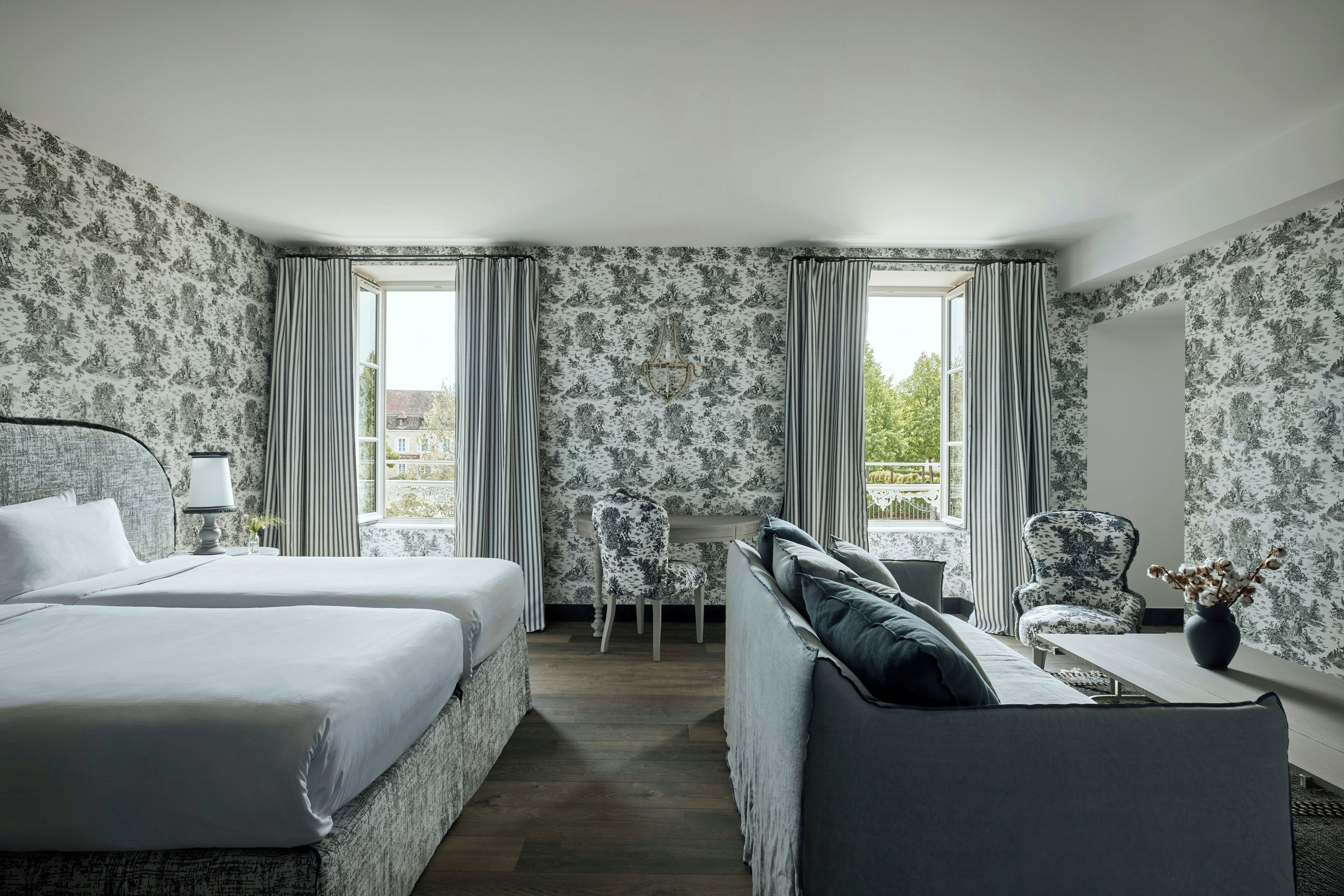 Een hotelkamer met een bed, een bank, een tafeltje en een stoel. Twee ramen met gordijnen en behang met een bloemenmotief.
