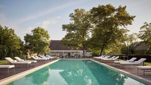 A rectangular swimming pool with lounge chairs on either side, surrounded by trees and a building in the background.