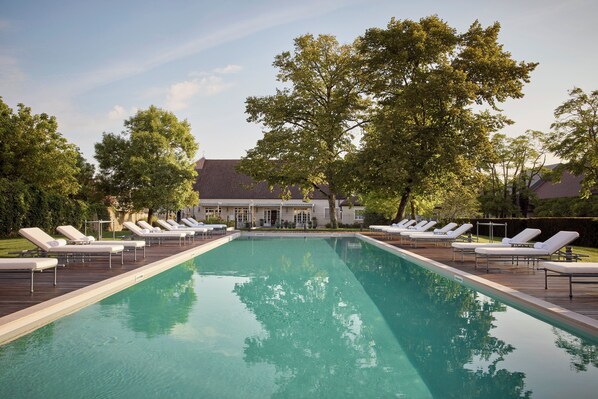 A rectangular swimming pool with lounge chairs on either side, surrounded by trees and a building in the background. - COMO Le Montrachet (Puligny-Montrachet)