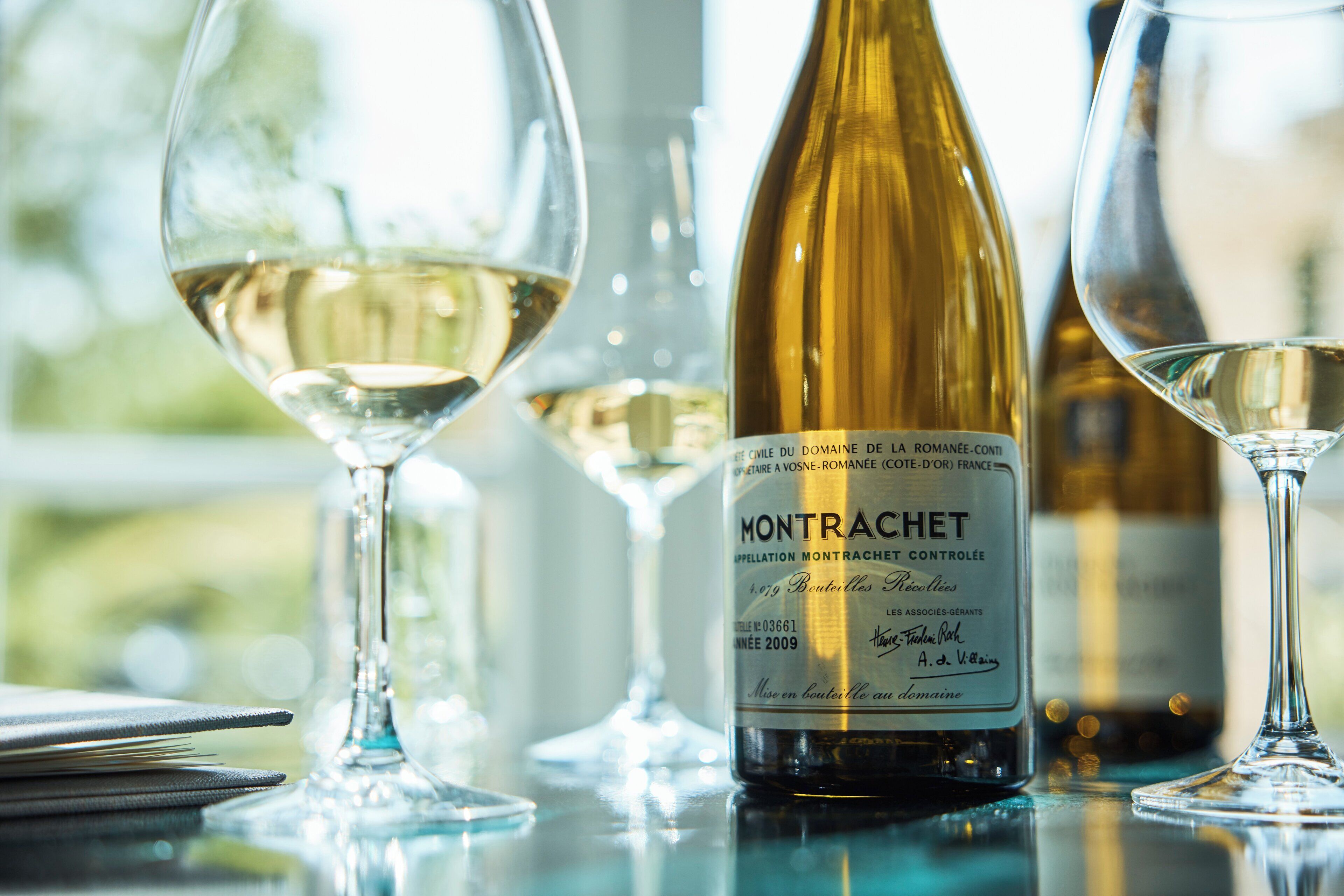 Une bouteille de vin de Montrachet, avec son étiquette, est posée sur une table, à côté de plusieurs verres de vin blanc.