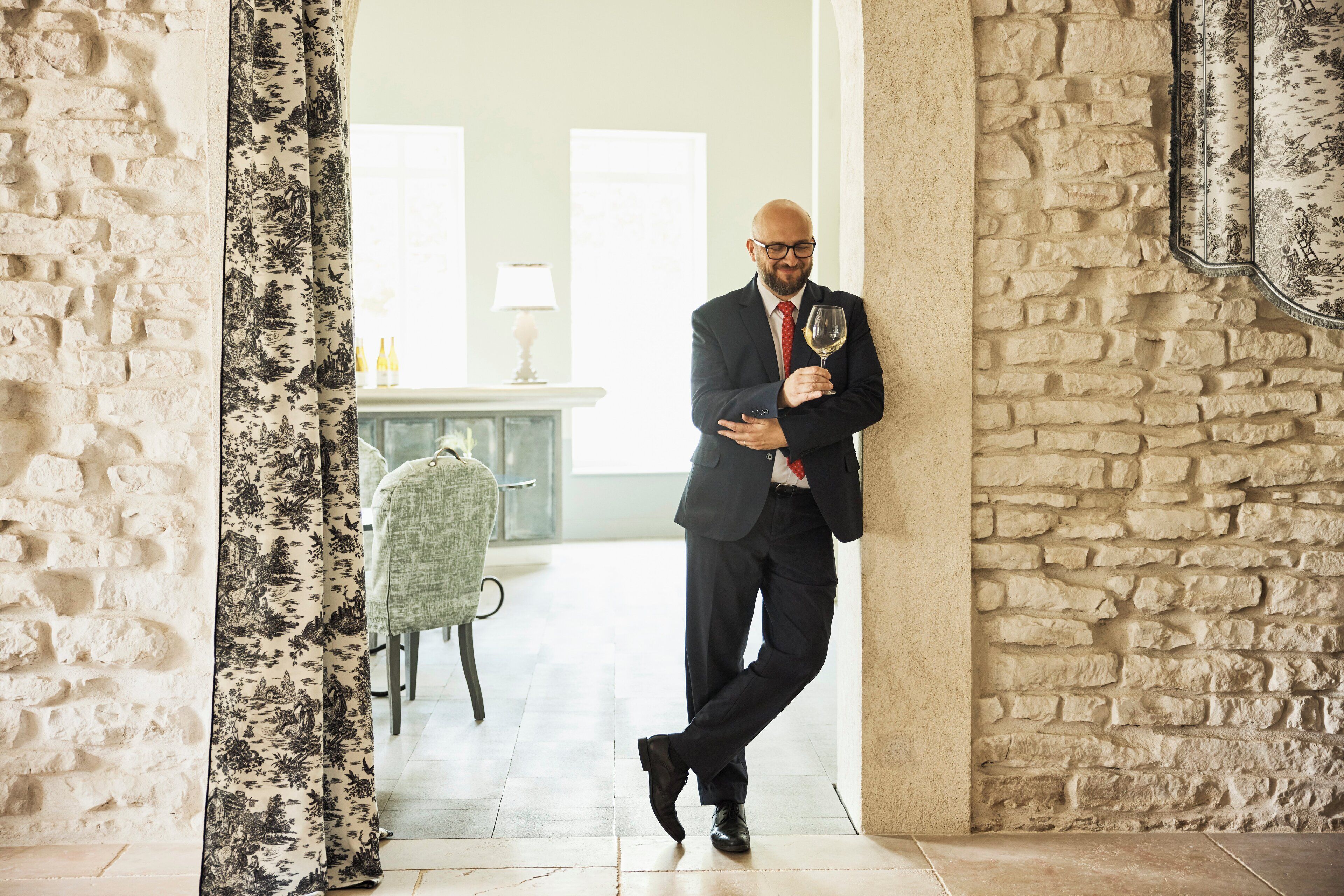 A man in a suit holding a wine glass, standing in a room with a stone wall and a patterned curtain.