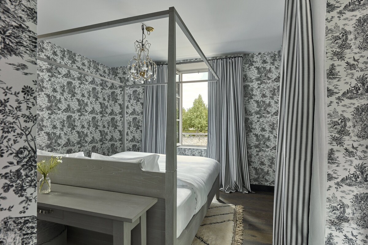 A bedroom with a four-poster bed, floral wallpaper, and a view of greenery outside.