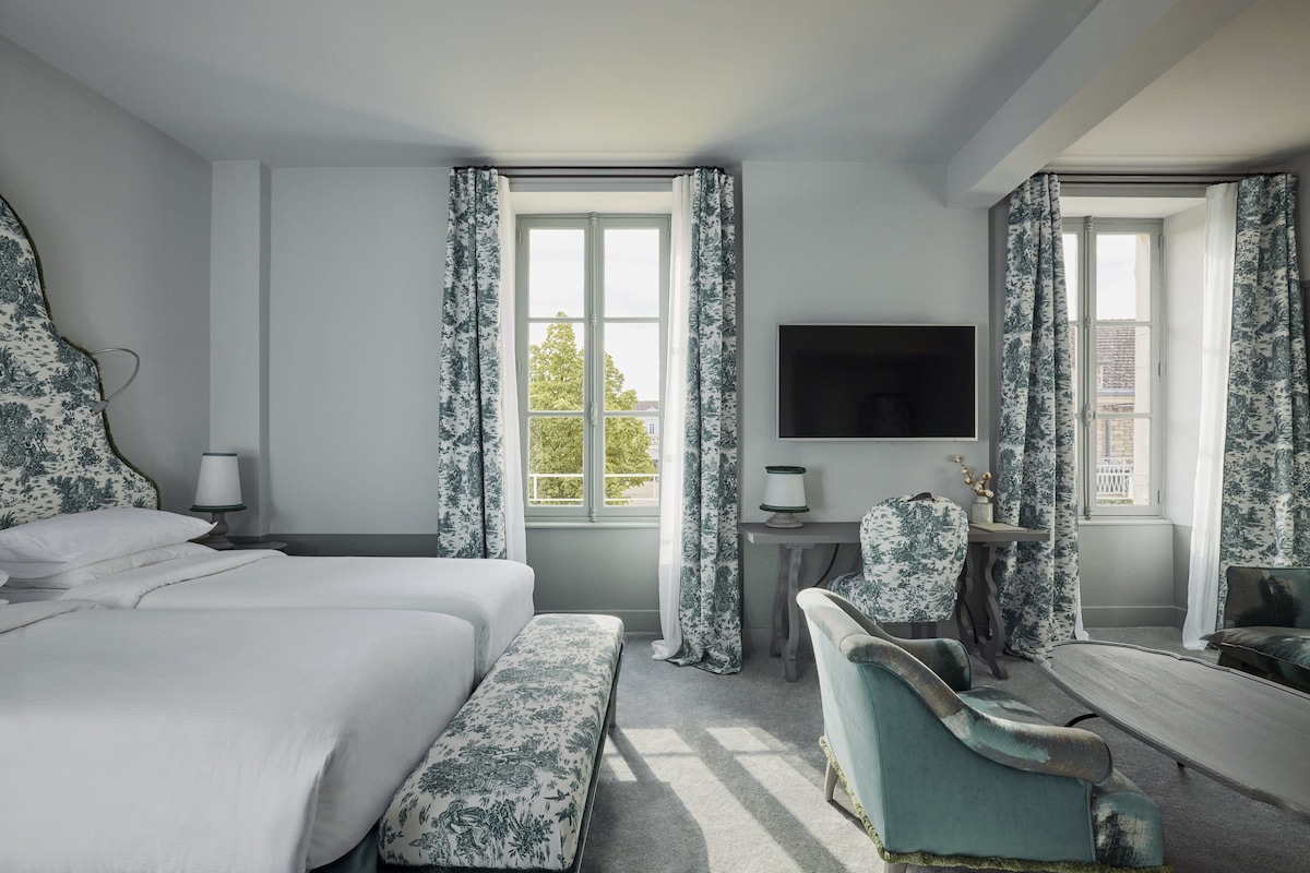 A hotel room with a large bed, a desk with a chair, a television, and two windows with curtains.