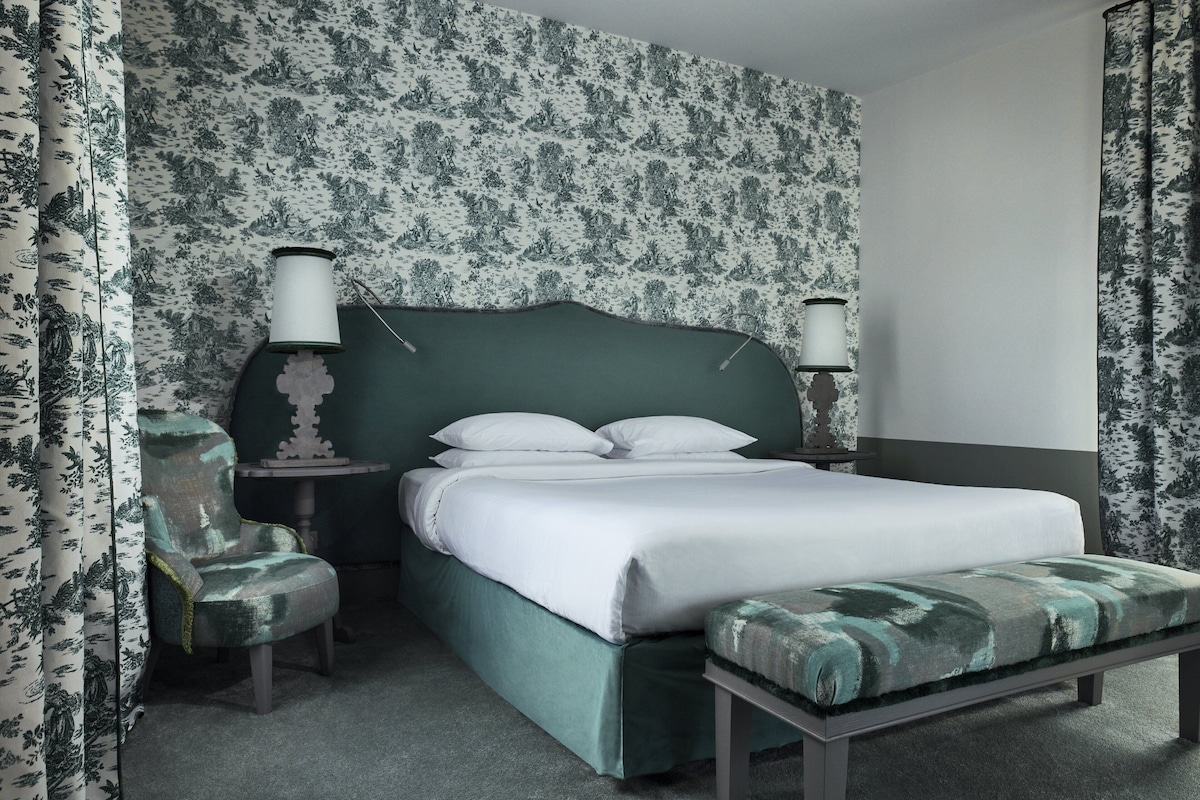 A bedroom with a green upholstered bed, two bedside lamps, a matching bench, and floral-patterned wallpaper.