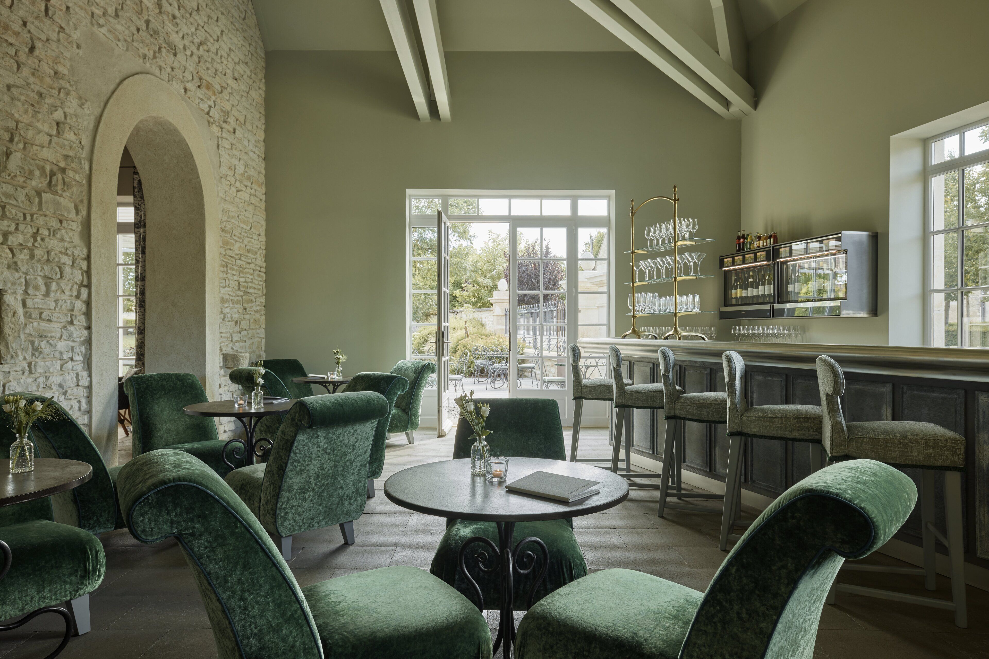 Un restaurant avec des chaises à revêtement vert, un mur en pierre et un bar doté d’étagères remplies de verres et de bouteilles.