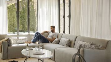 A man sitting on a sofa in a well-lit room with a large window, a round coffee table, and a remote control on the floor.