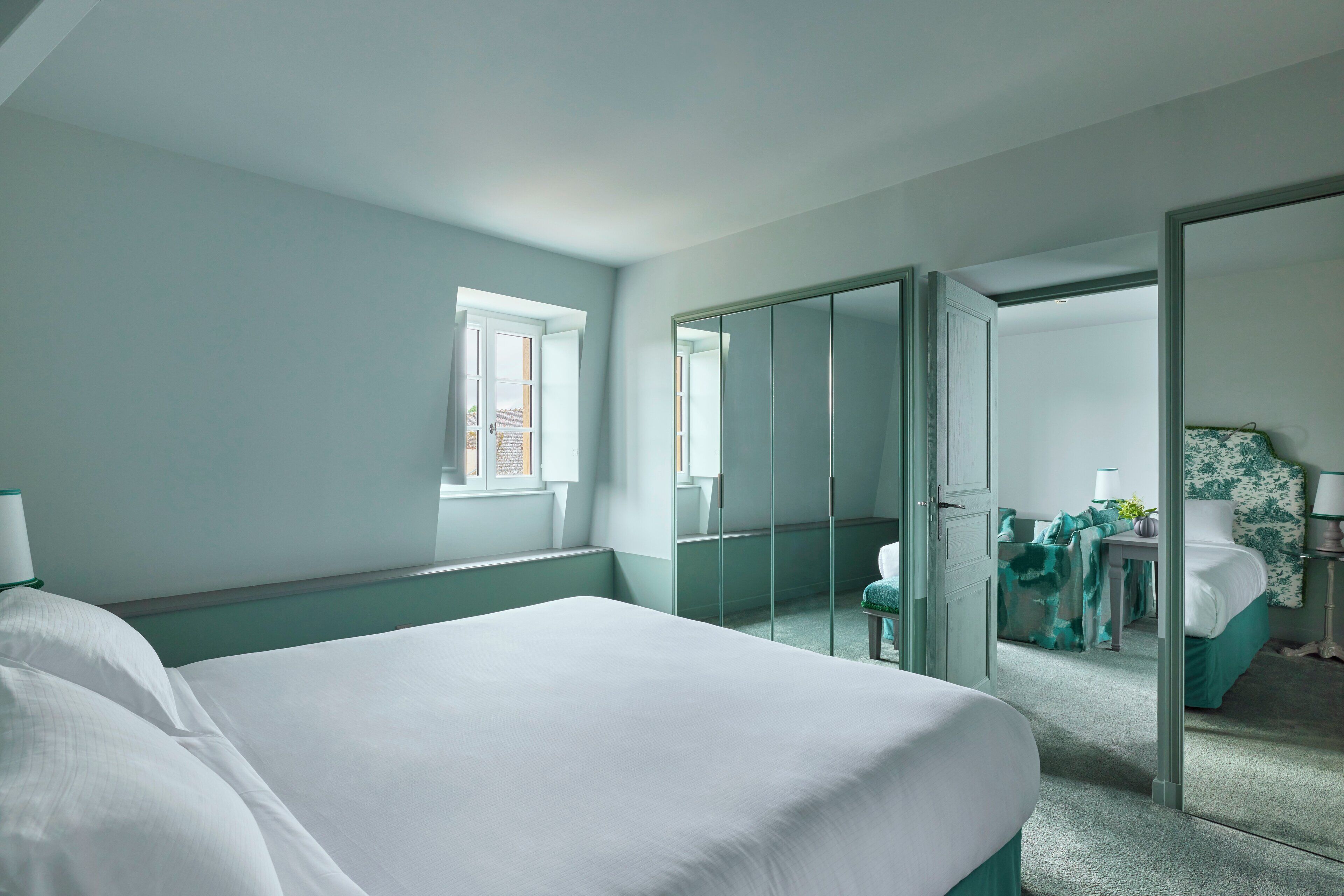 Quarto de hotel com uma cama grande, um espelho, uma janela e uma porta que dá para outro cômodo.