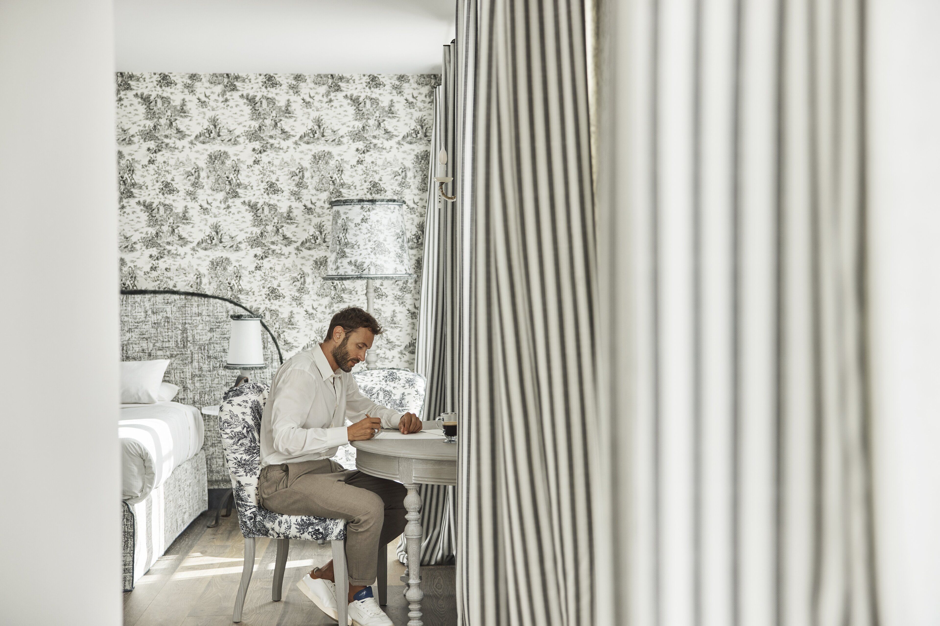 A man sitting at a table in a room with a patterned wallpaper, a bed, and a lamp.