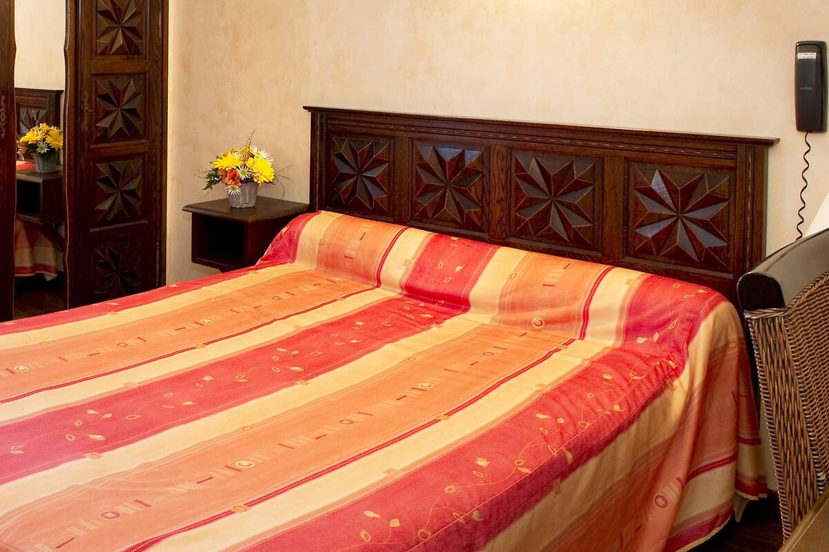 A hotel room with a bed, a nightstand, a vase with flowers, and a telephone on the wall.