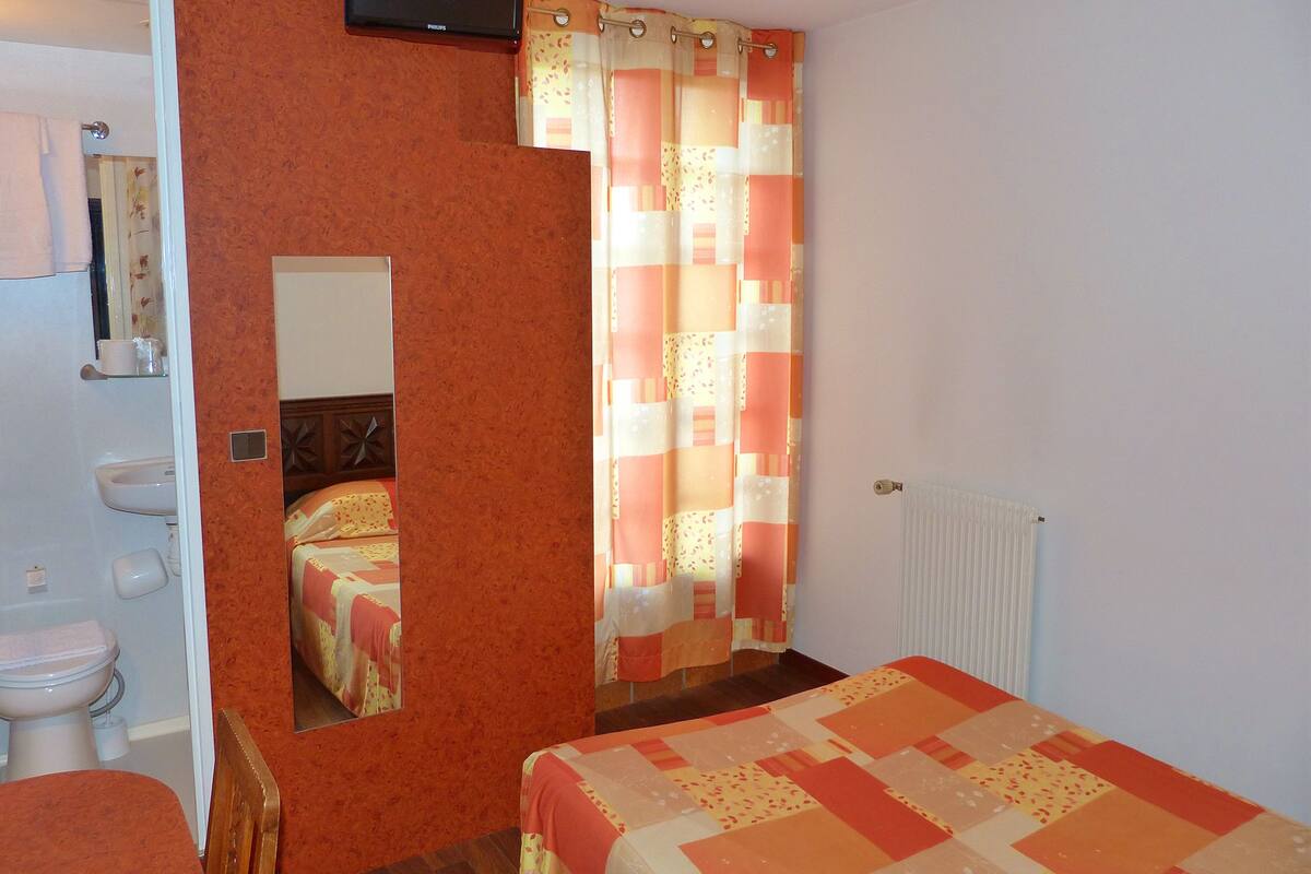 A bedroom with a bed, a mirror, a television, a bathroom with a toilet and sink, and a window with curtains.