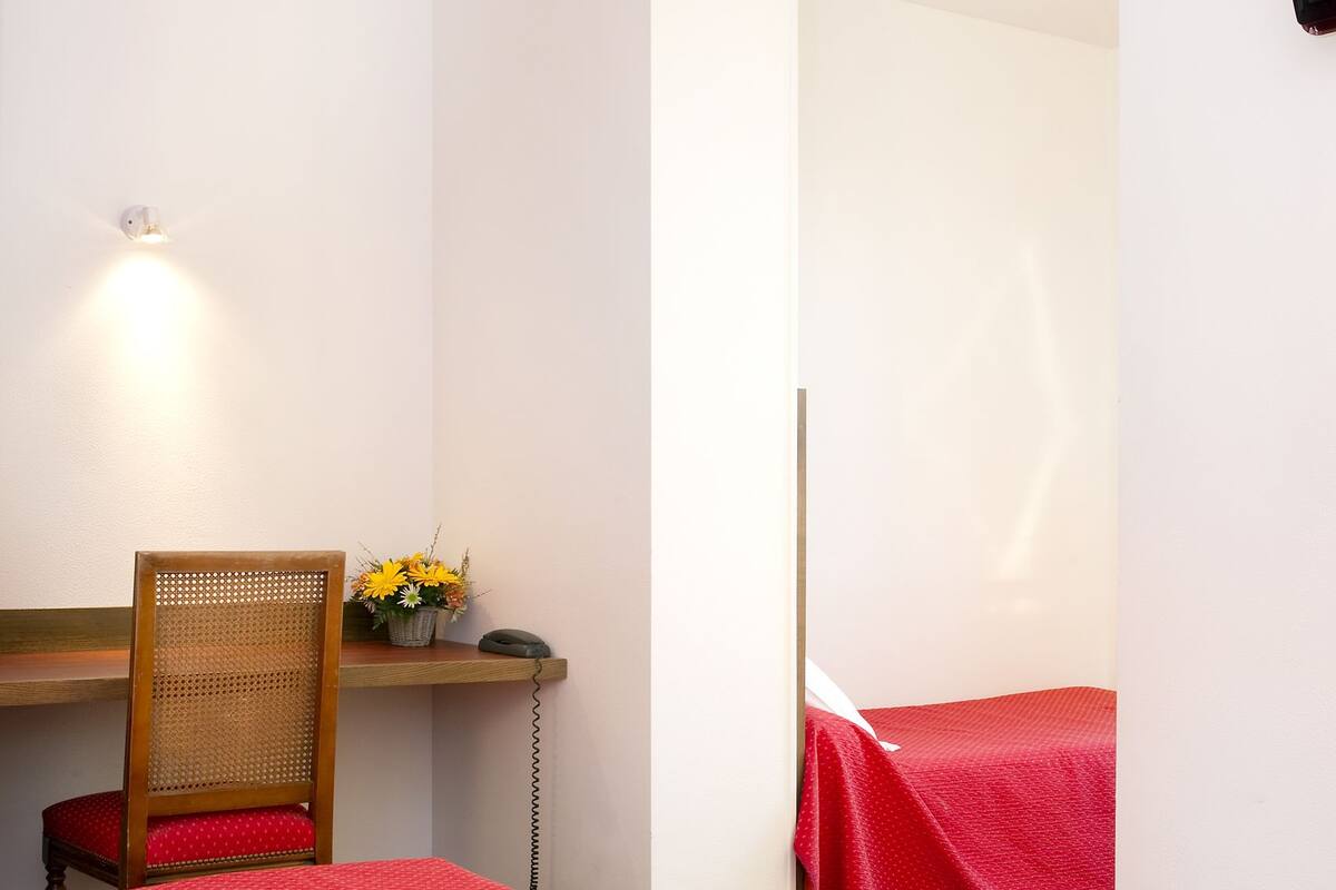 A small, bright room with two red beds, a wooden chair, a desk with a lamp, and a wall-mounted TV.