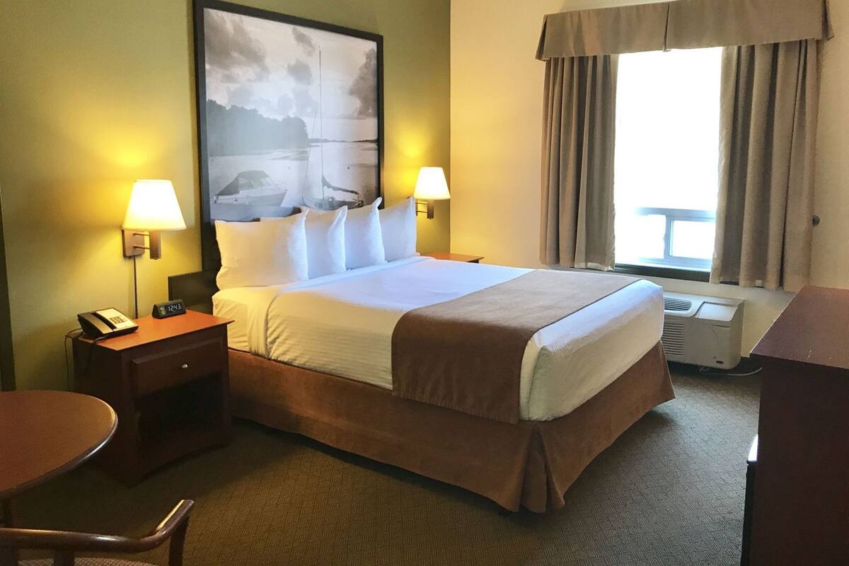 A hotel room with a bed, two bedside lamps, a desk with a telephone, a chair, a window with curtains, and a wall-mounted air conditioner.