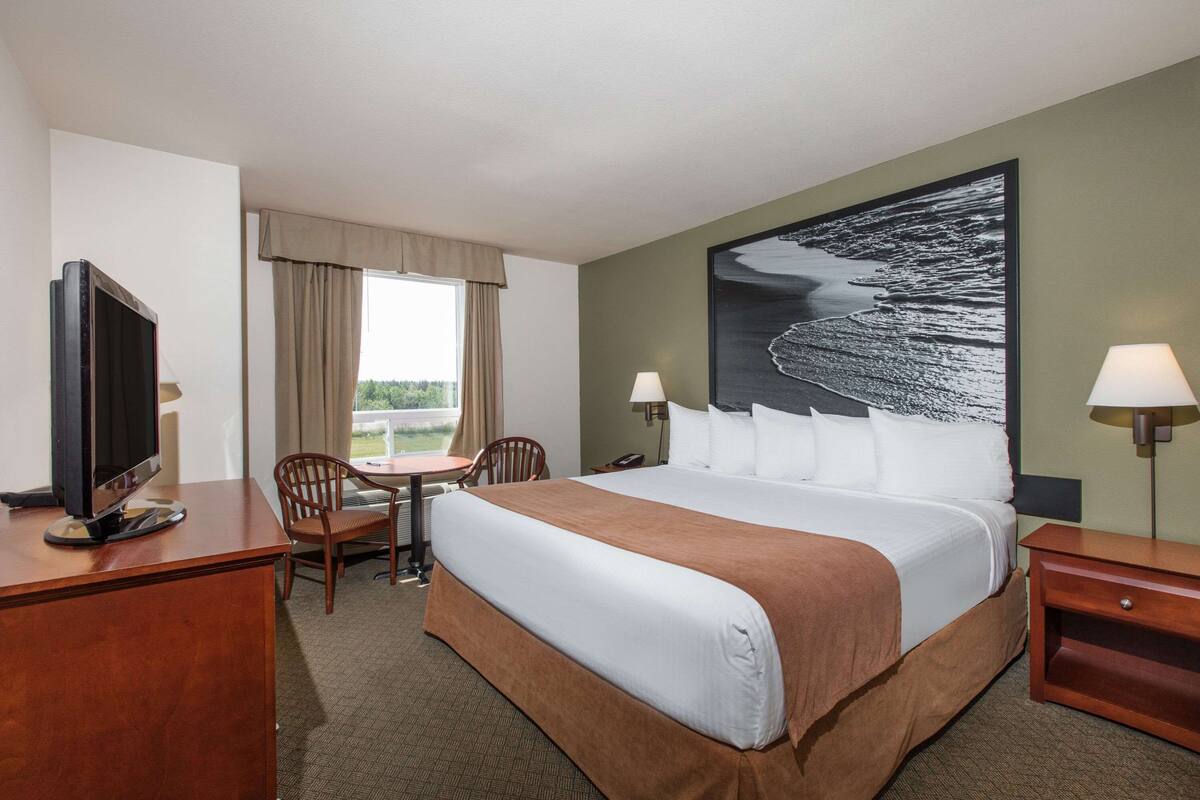 A hotel room with a large bed, a desk with a television, two chairs, a window with curtains, and a painting on the wall.