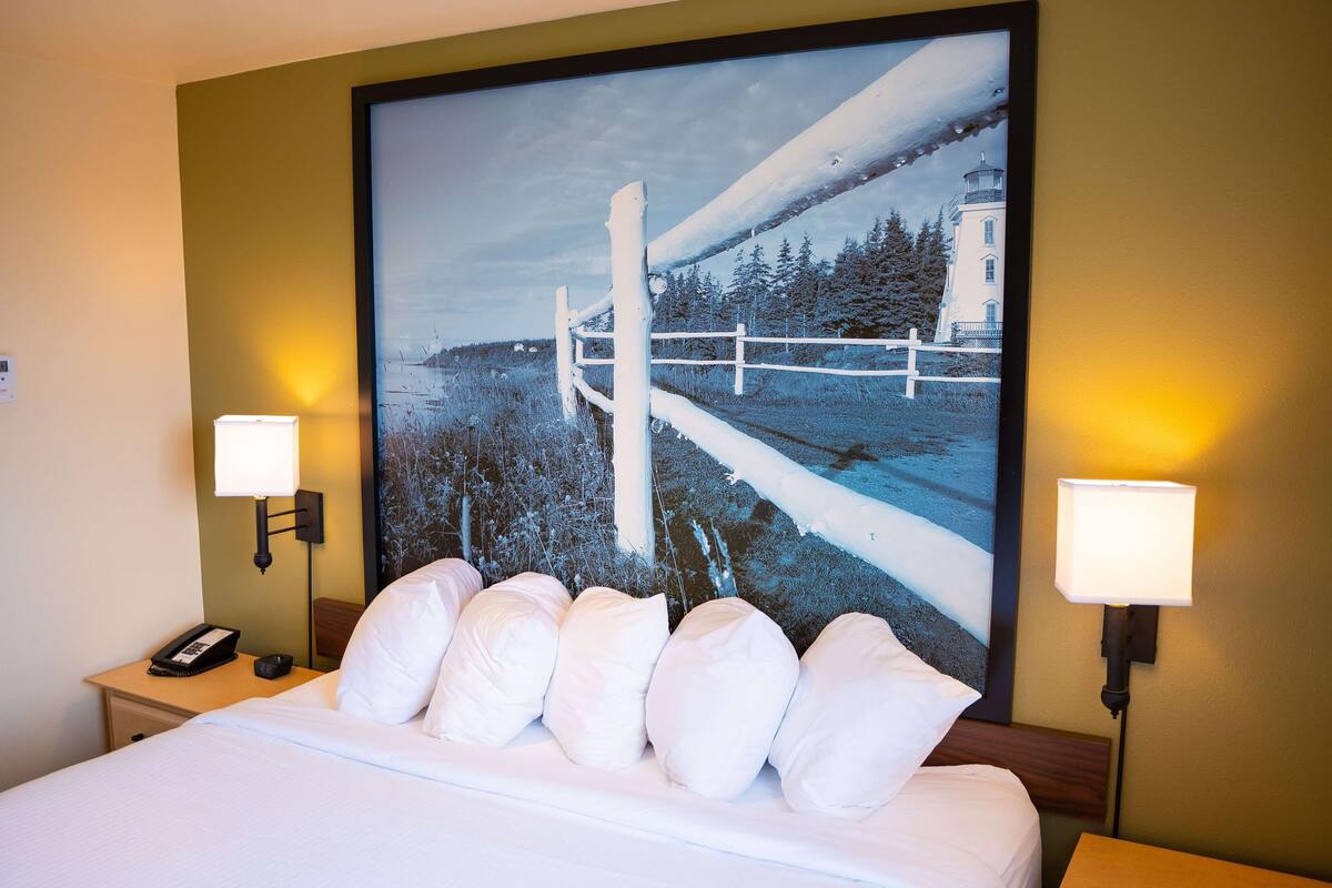 A hotel room with a large bed, white bedding, two bedside lamps, a nightstand with a phone, and a painting of a lighthouse on the wall.