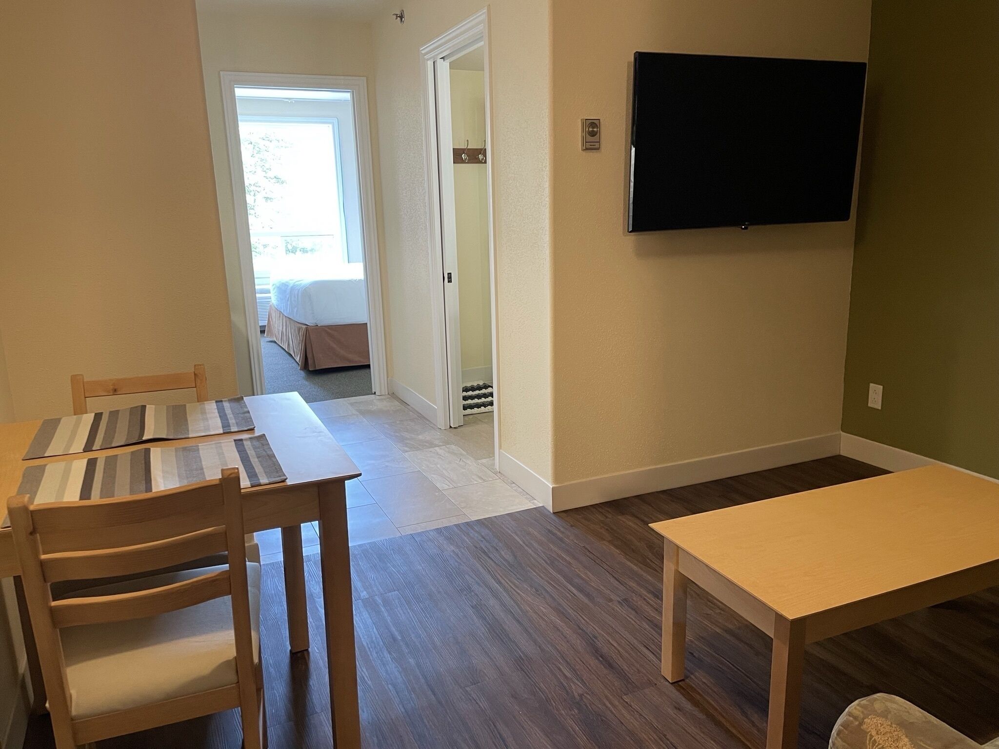 Een kamer met een houten eettafel en stoelen, een wandgemonteerde flatscreen televisie, en uitzicht op de aangrenzende slaapkamer.