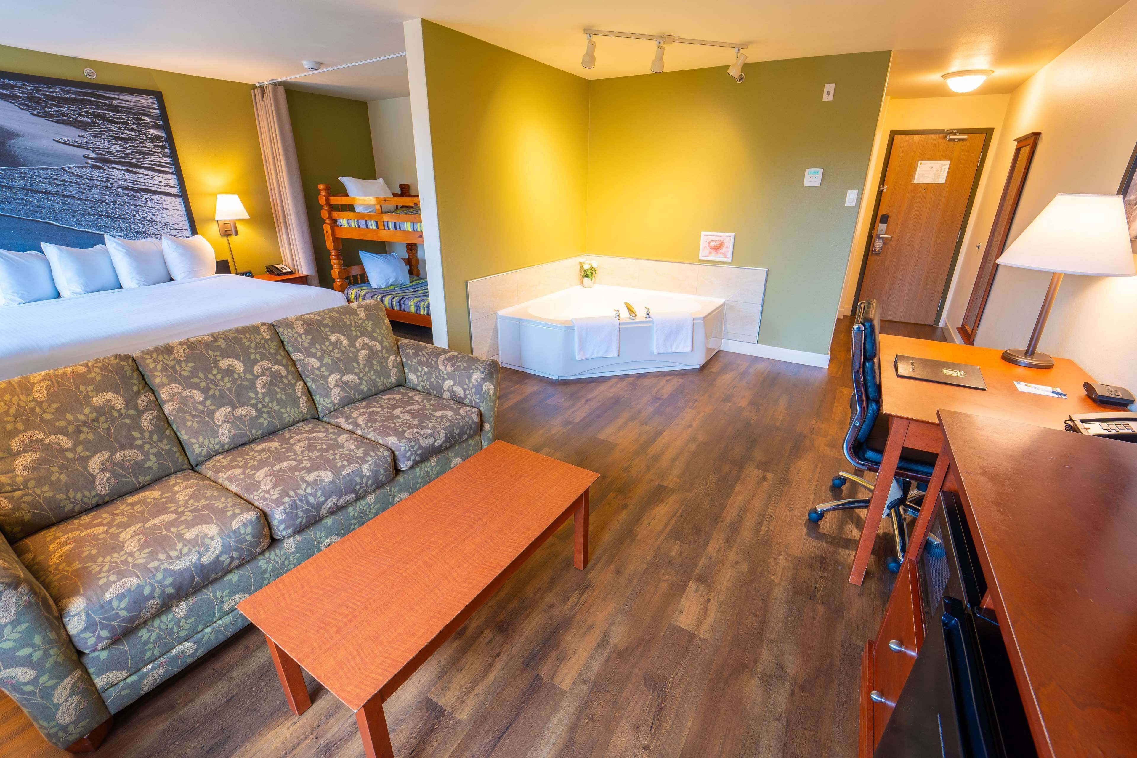A hotel room with a bed, sofa, wooden table, desk, and a bathtub.