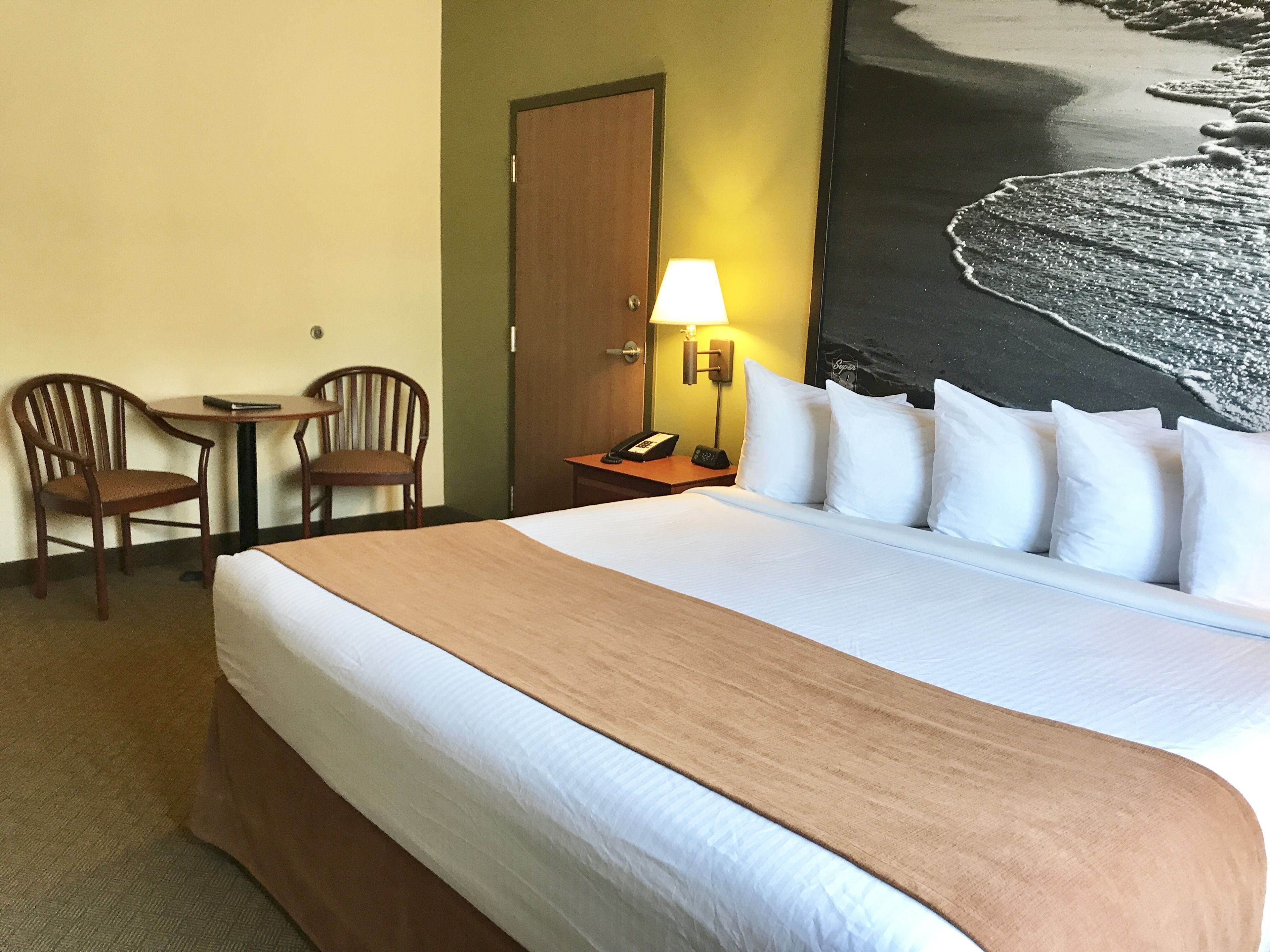 A hotel room with a large bed, two chairs, a small table, a lamp, and a painting of a beach scene.
