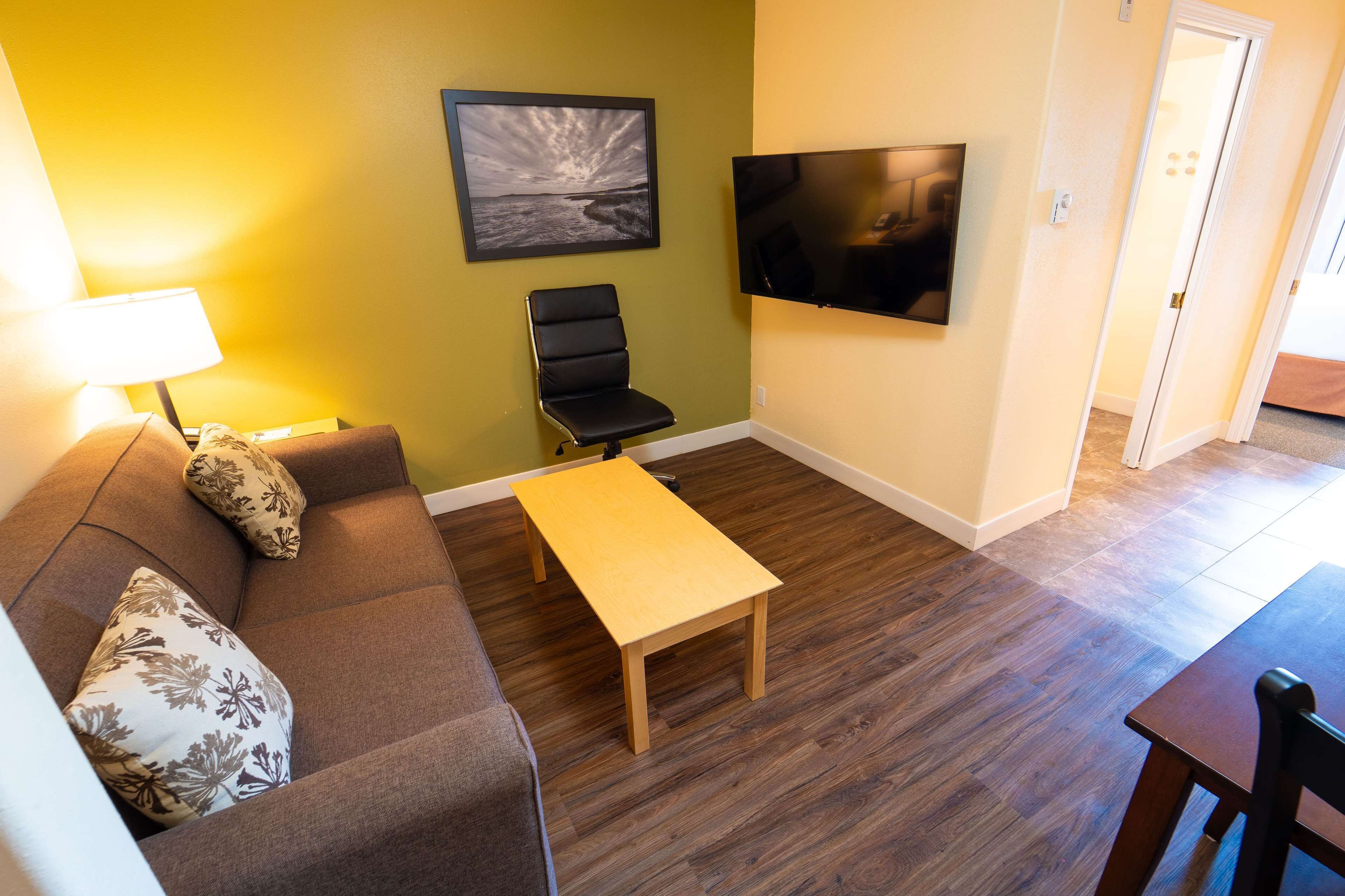 En stue med sofa, et lille bord, en sort stol, et fjernsyn ophængt på væggen og en dør, der fører til et andet rum.