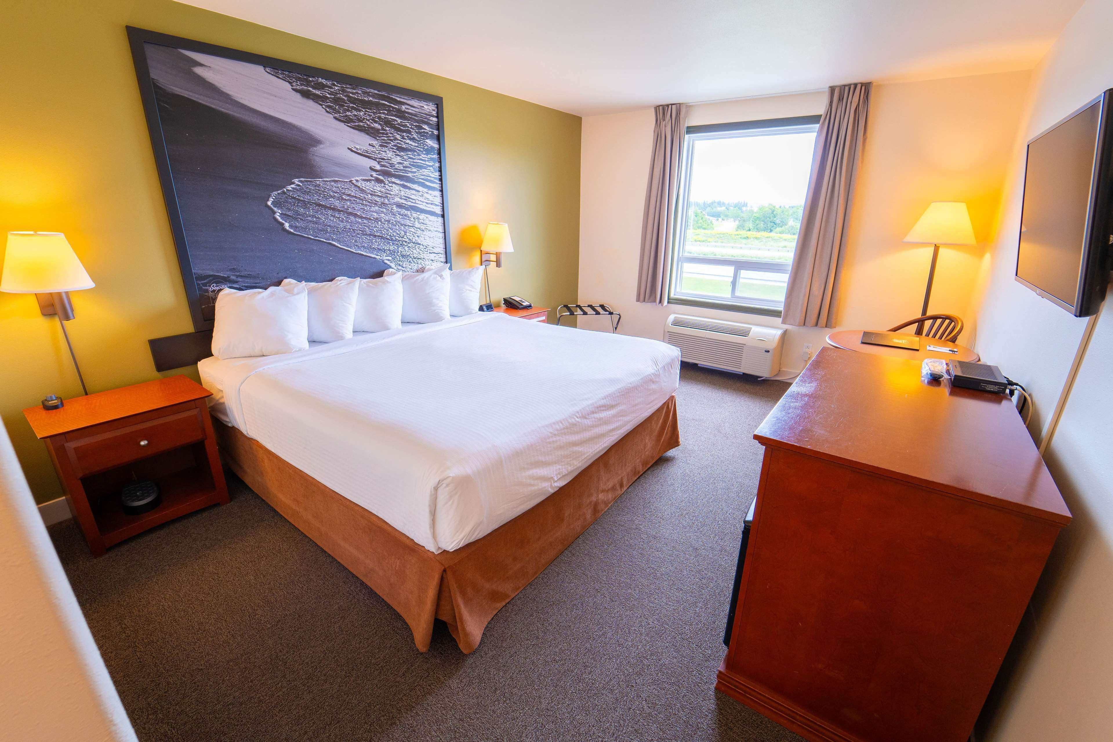 A hotel room with a large bed, two bedside tables with lamps, a wooden dresser, a flat-screen TV, and a large window with curtains.
