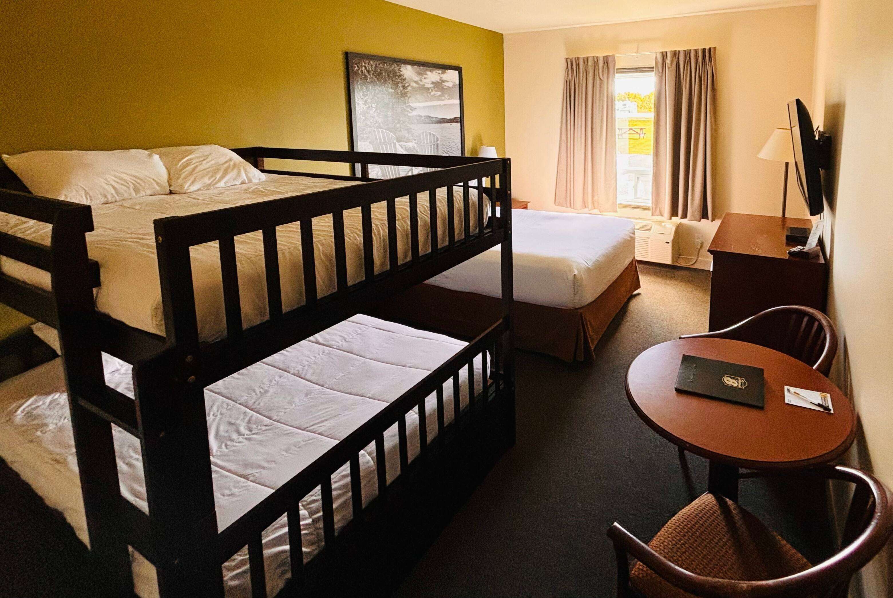 Quarto de hotel com cama beliche, cama de solteiro, escrivaninha com cadeira, televisão e janela com cortinas.