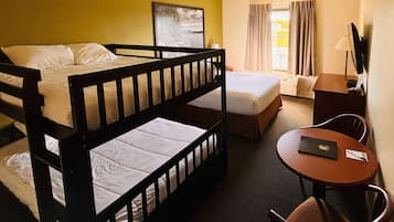 Quarto de hotel com cama beliche, cama de solteiro, escrivaninha com cadeira, televisão e janela com cortinas.