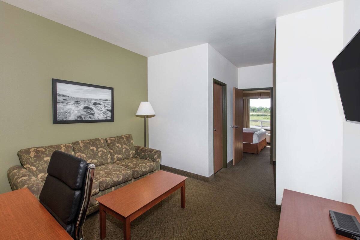 A hotel room with a sofa, a wooden table, a lamp, a framed picture on the wall, and a view of the outdoors through a door.