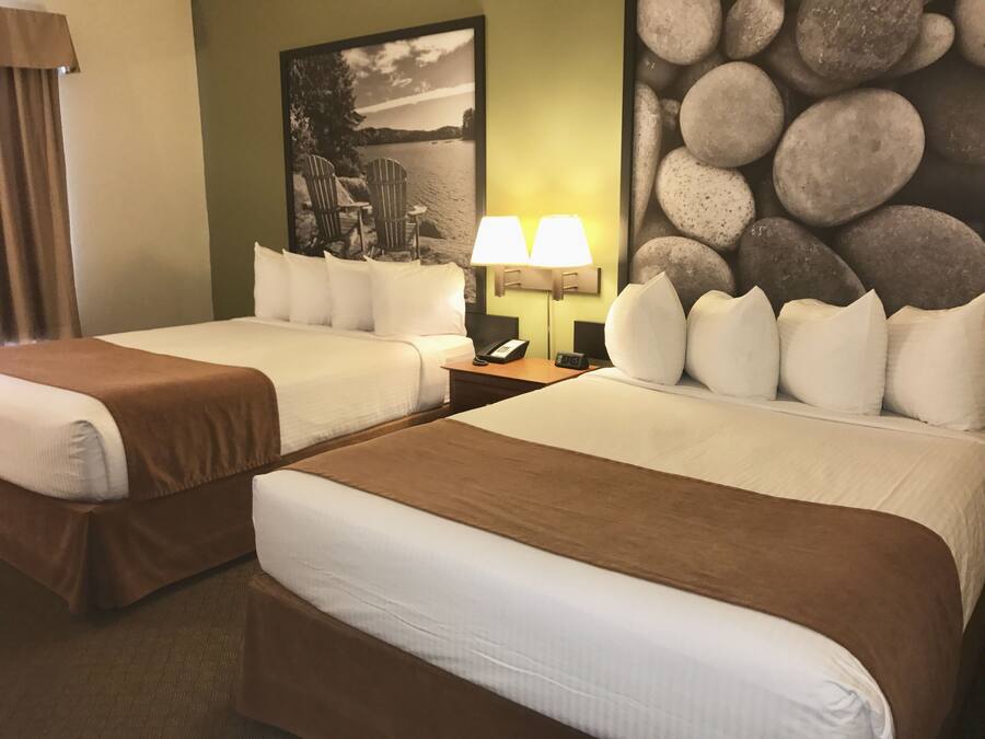 Habitación de hotel con dos camas, una mesita de noche con lámparas, un cuadro con sillas junto a un lago y una pared de piedra.