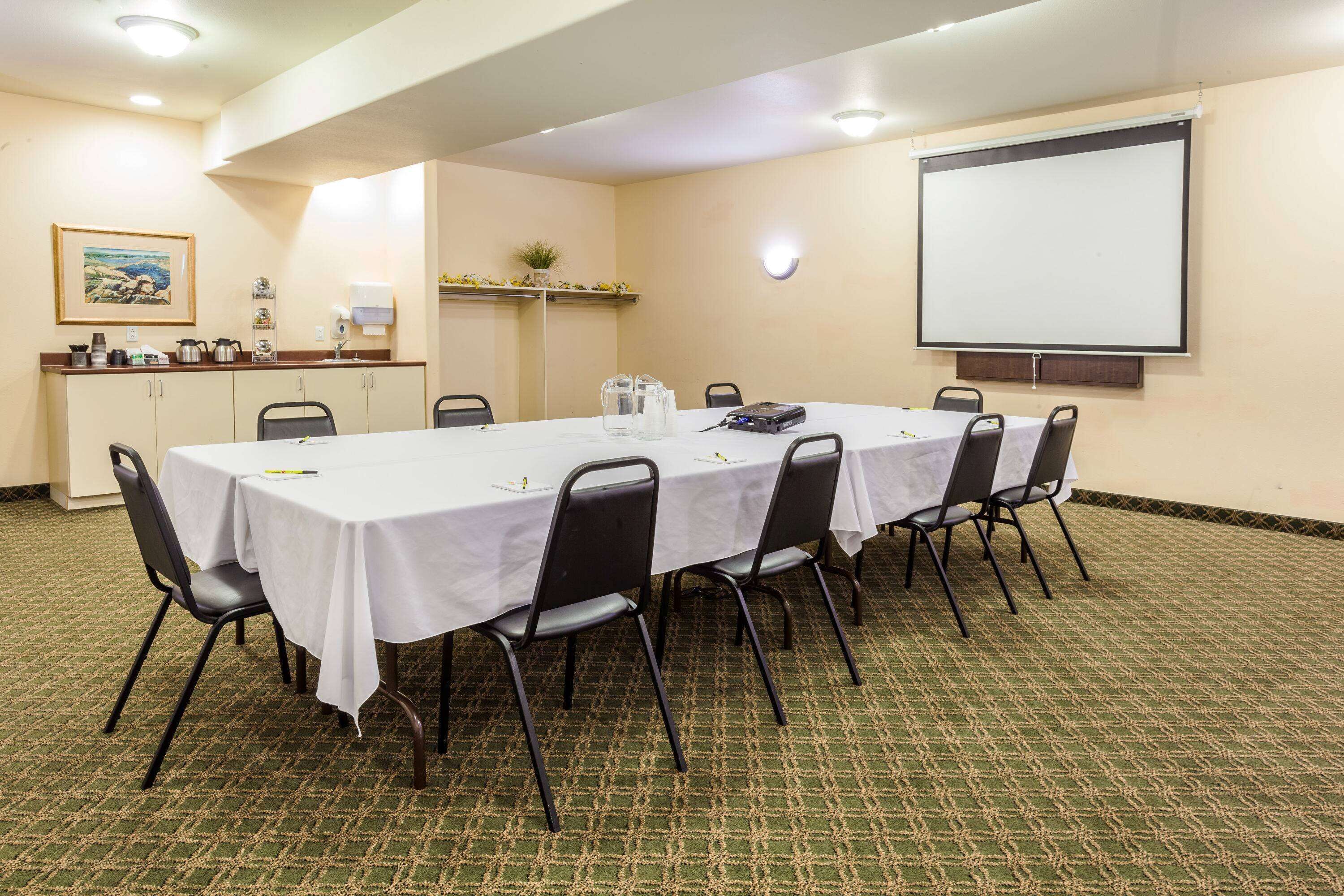 Sala de conferências com uma mesa comprida, cadeiras, tela de projetor e uma pequena área de copa.