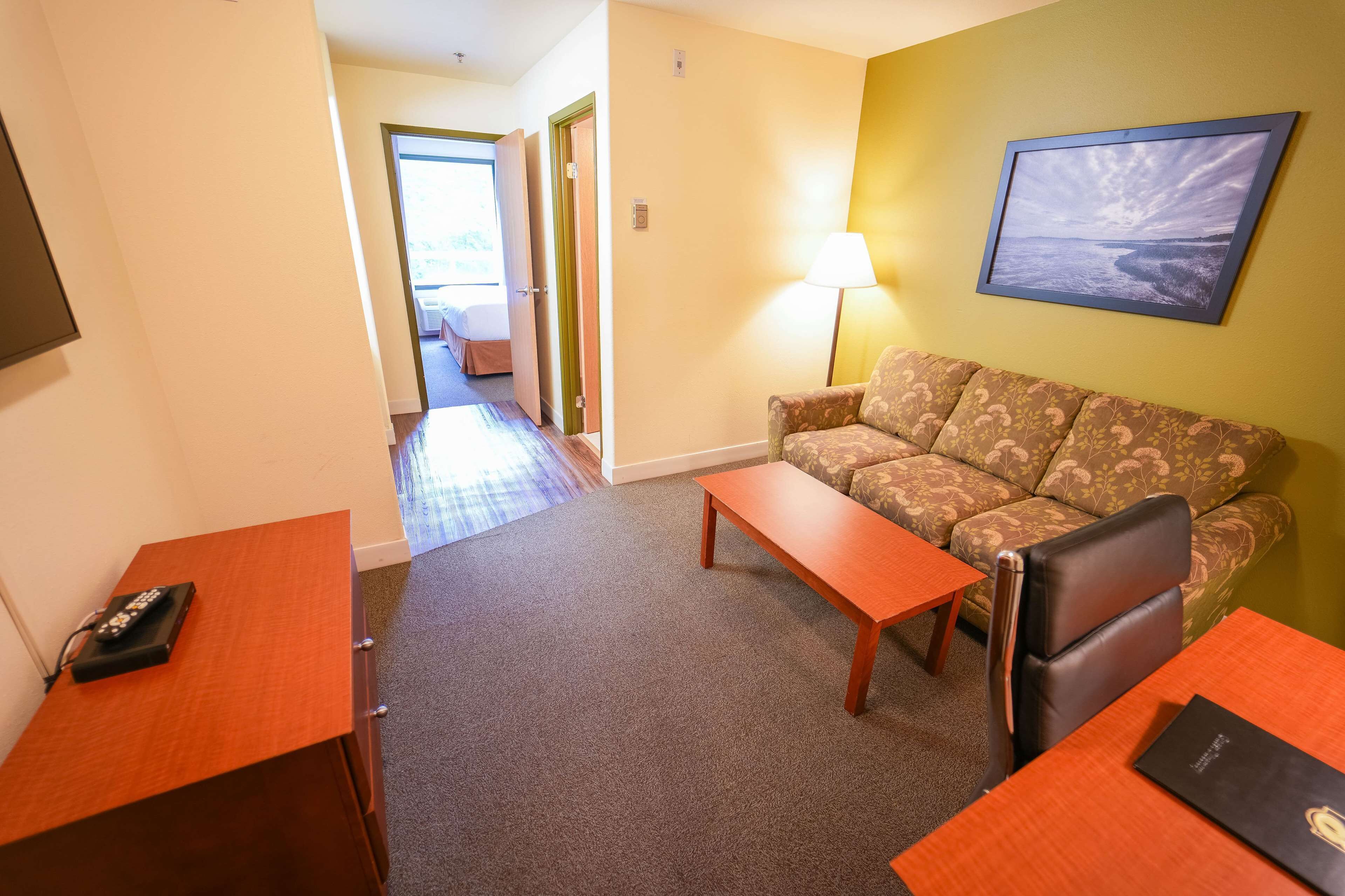 A hotel room with a sofa, a coffee table, a desk, and a lamp. There is a framed picture on the wall and a door leading to another room.