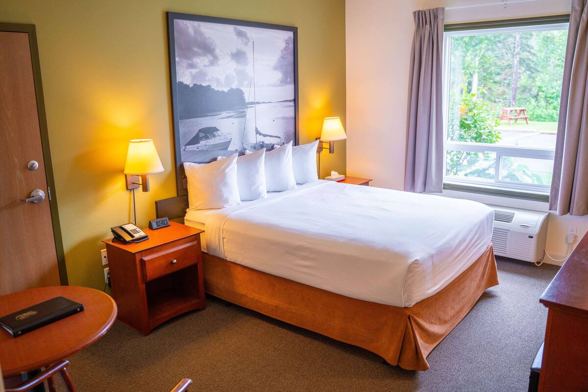 A hotel room with a large bed, two bedside lamps, a nightstand with a phone, a small table with a book, and a window with curtains.