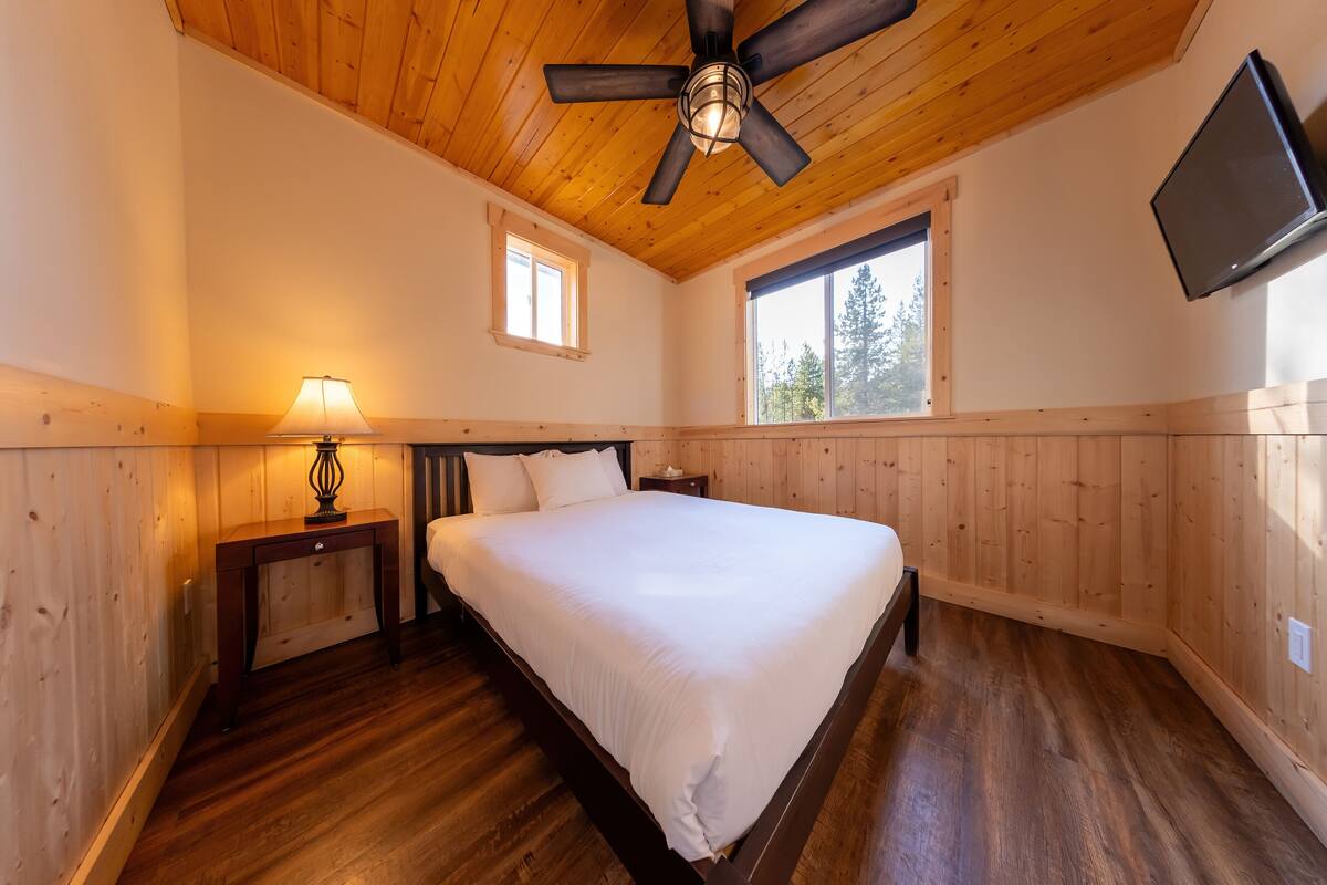 A bedroom with a wooden ceiling, a bed, a nightstand with a lamp, and a TV mounted on the wall.