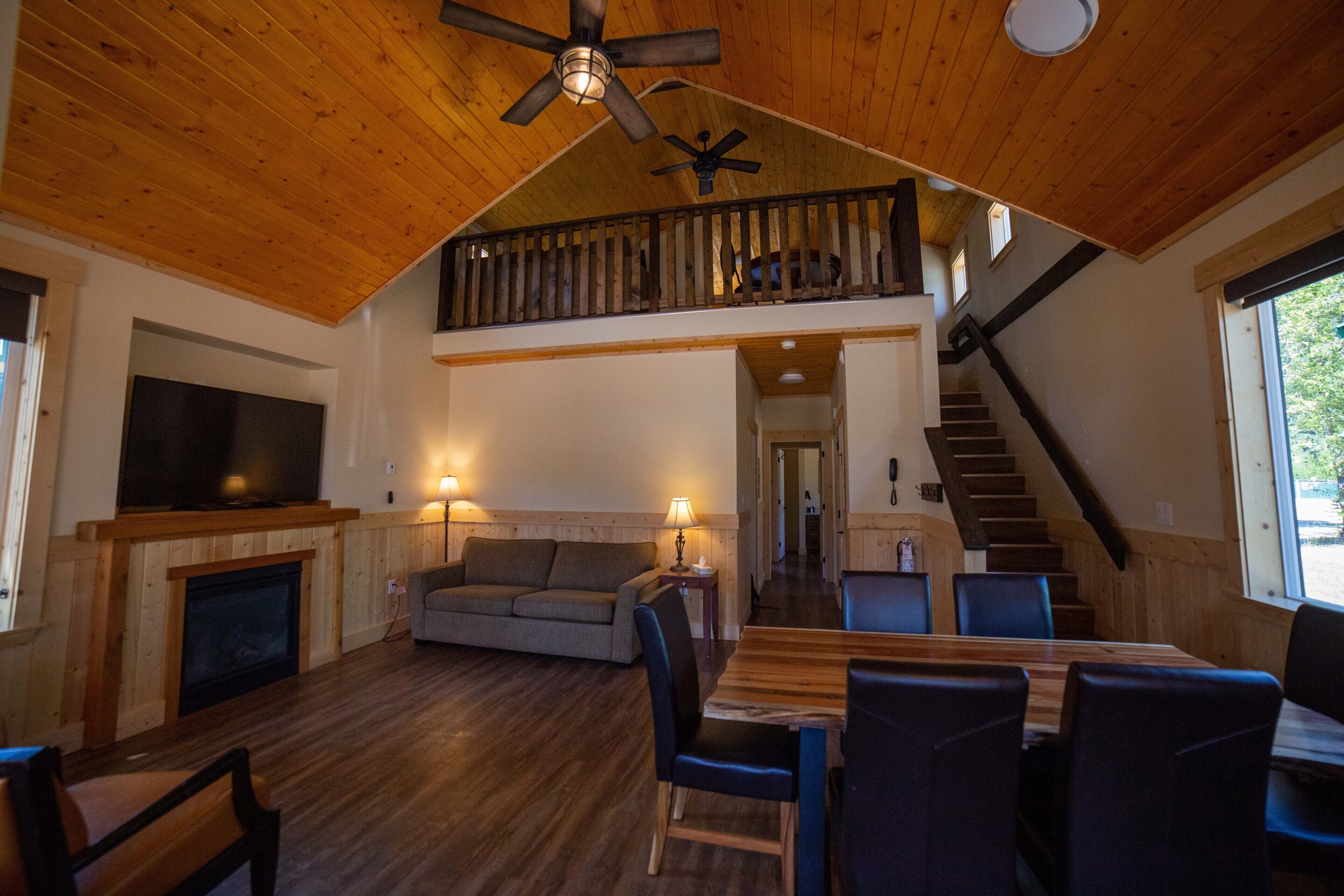 A spacious living area with a fireplace, a sofa, a dining table with chairs, a TV mounted on the wall, and a staircase leading to an upper level.