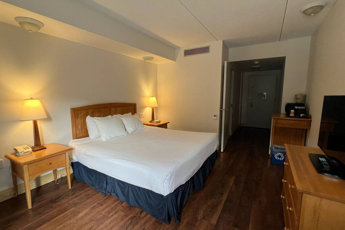 A hotel room with a large bed, two bedside lamps, a wooden nightstand, a desk with a telephone, and a television on a wooden stand.