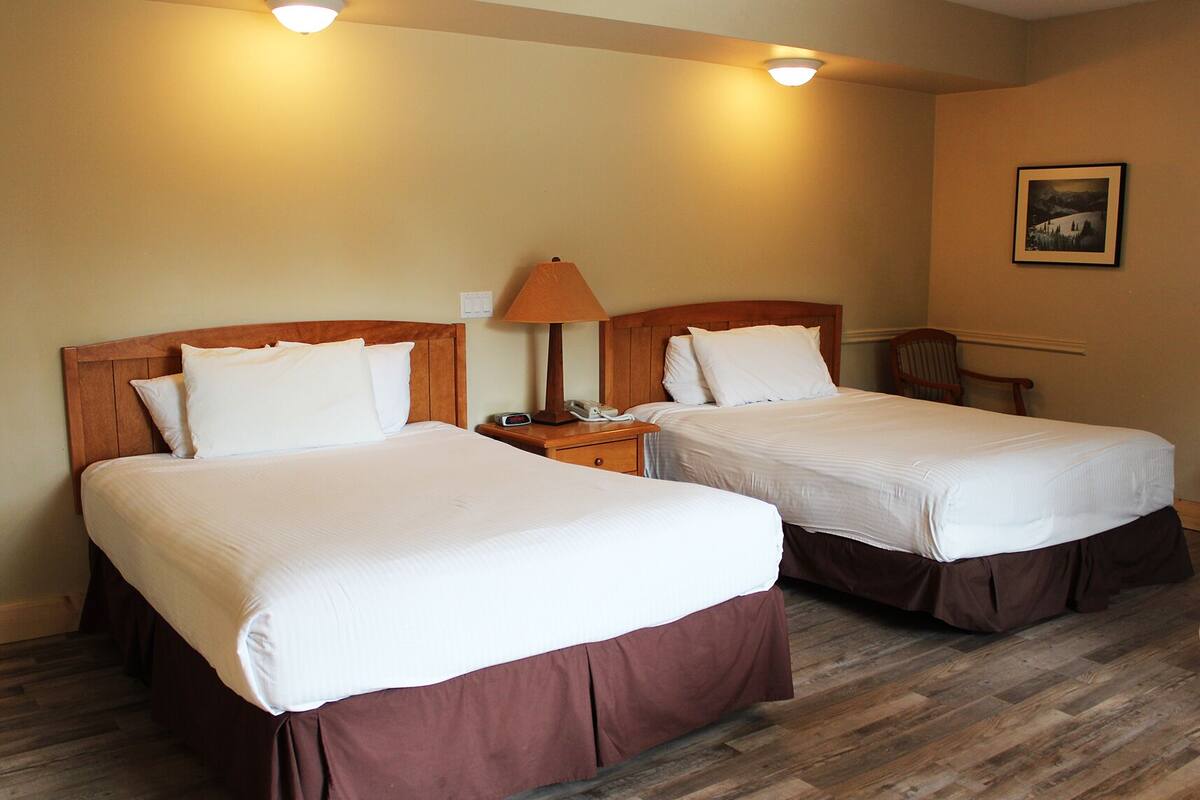 A hotel room with two beds, wooden headboards, a nightstand with a lamp, and a framed picture on the wall.