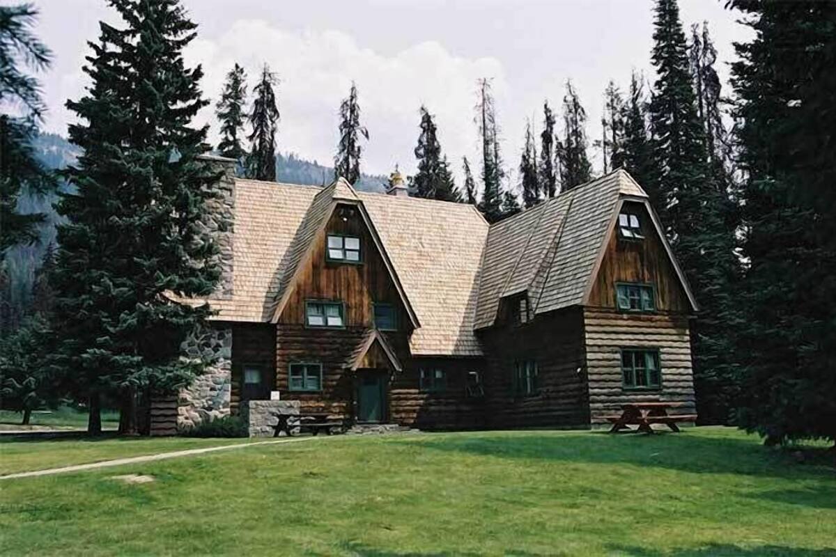 A log cabin with a symmetrical design, featuring a central entrance and two side wings, surrounded by a forested landscape.