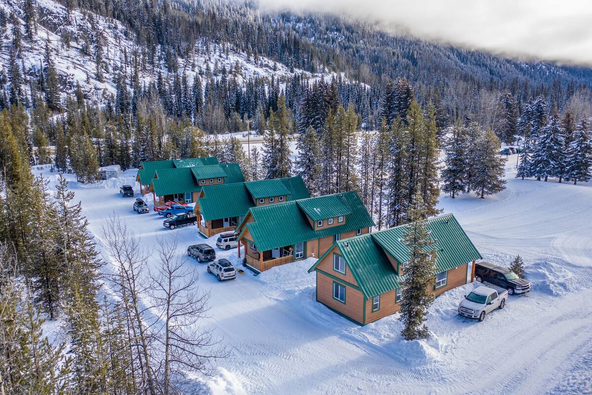 A snow-covered lodge with multiple green-roofed cabins, a parking area, and a forested mountain backdrop.