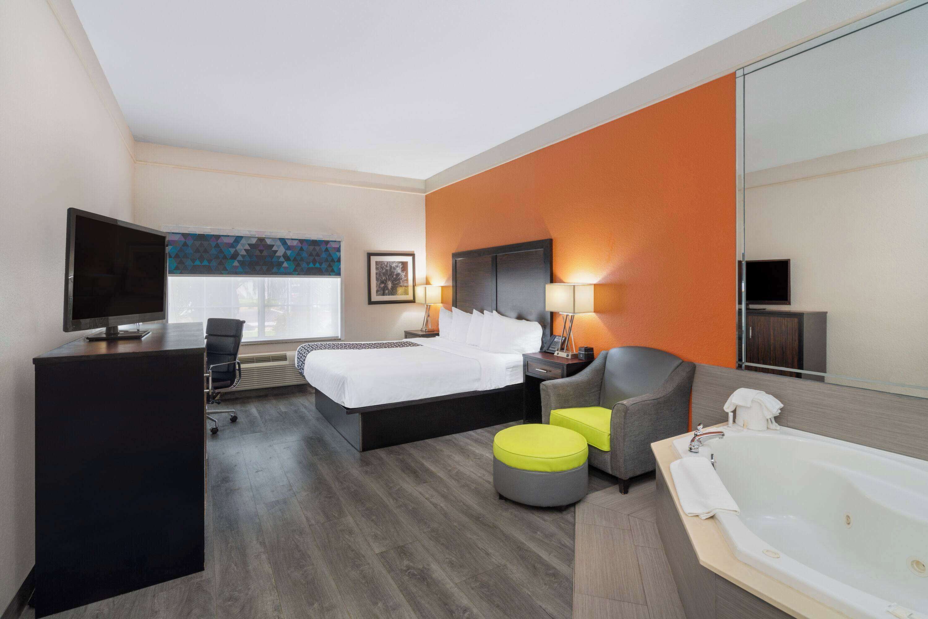 A modern hotel room with a bed, desk, chair, ottoman, and a large bathtub.