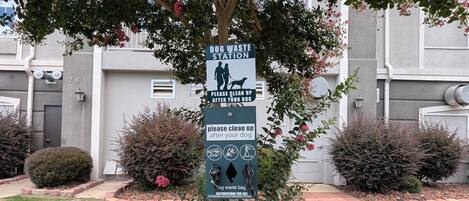 Estación para depositar desechos de perro con un letrero que indica limpiar después de su mascota, ubicada en una zona residencial con un árbol, arbustos y un edificio al fondo.