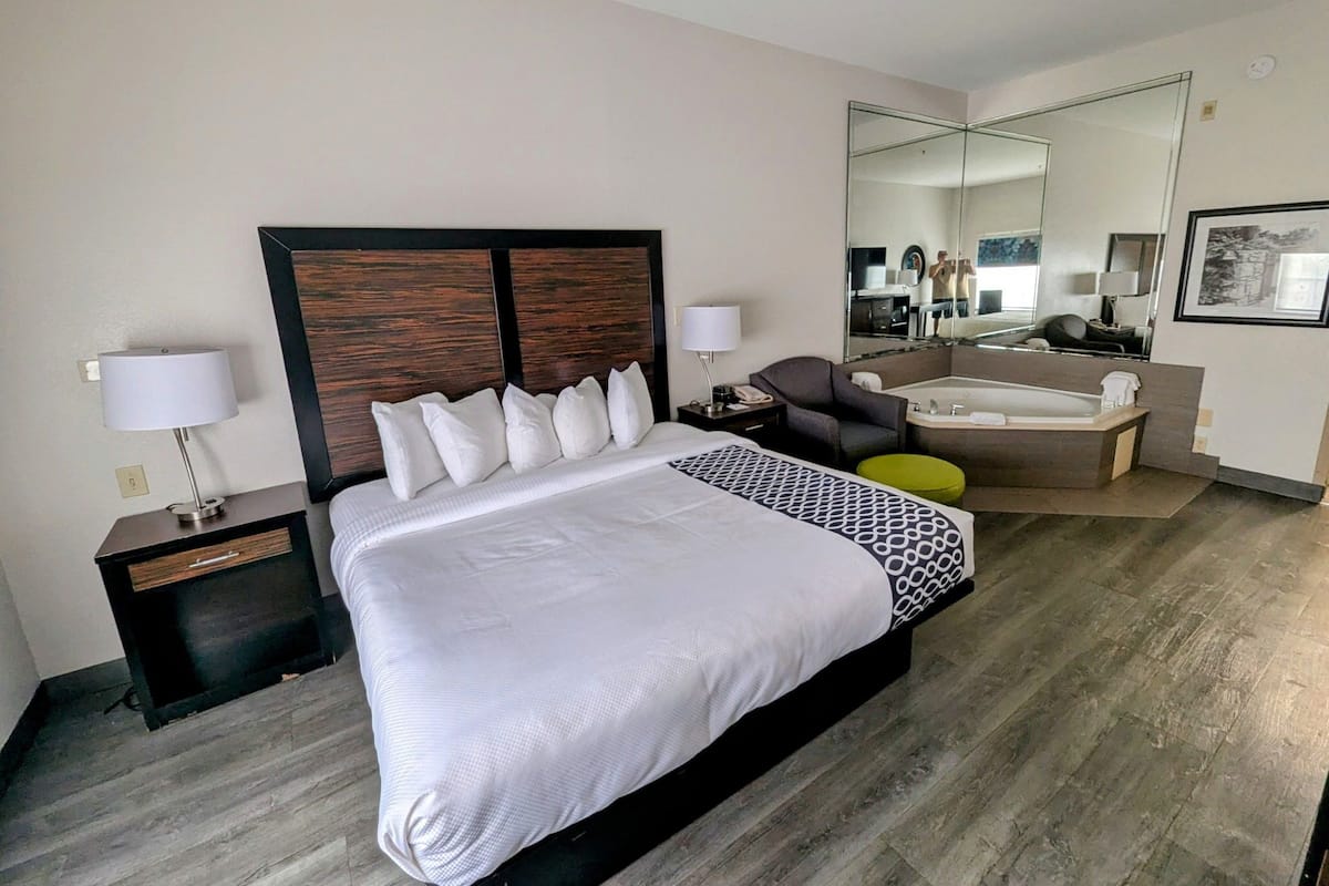 Une chambre d’hôtel avec un grand lit, une table de chevet avec une lampe, une chaise, une petite table ronde, un miroir et une salle de bain avec une baignoire.