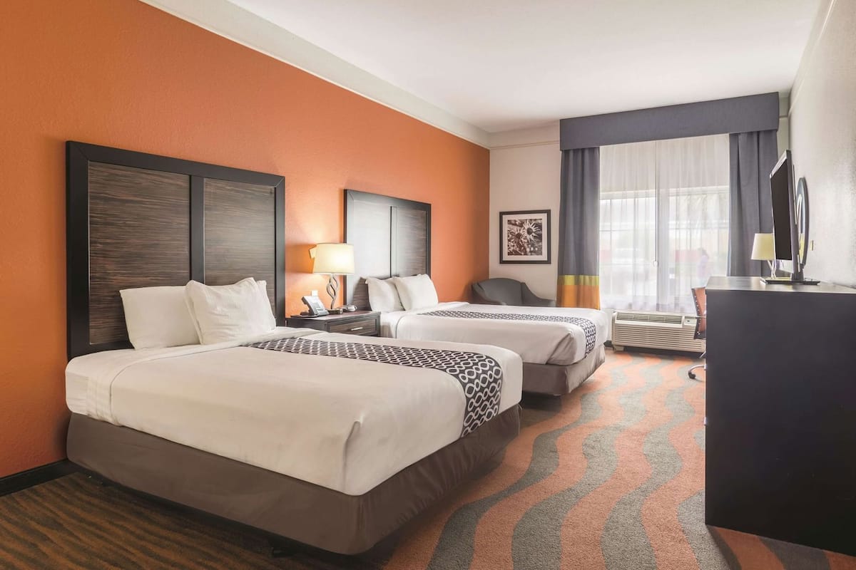 Une chambre d’hôtel avec deux lits, un bureau, une chaise, une lampe, un tableau au mur et une fenêtre avec des rideaux.